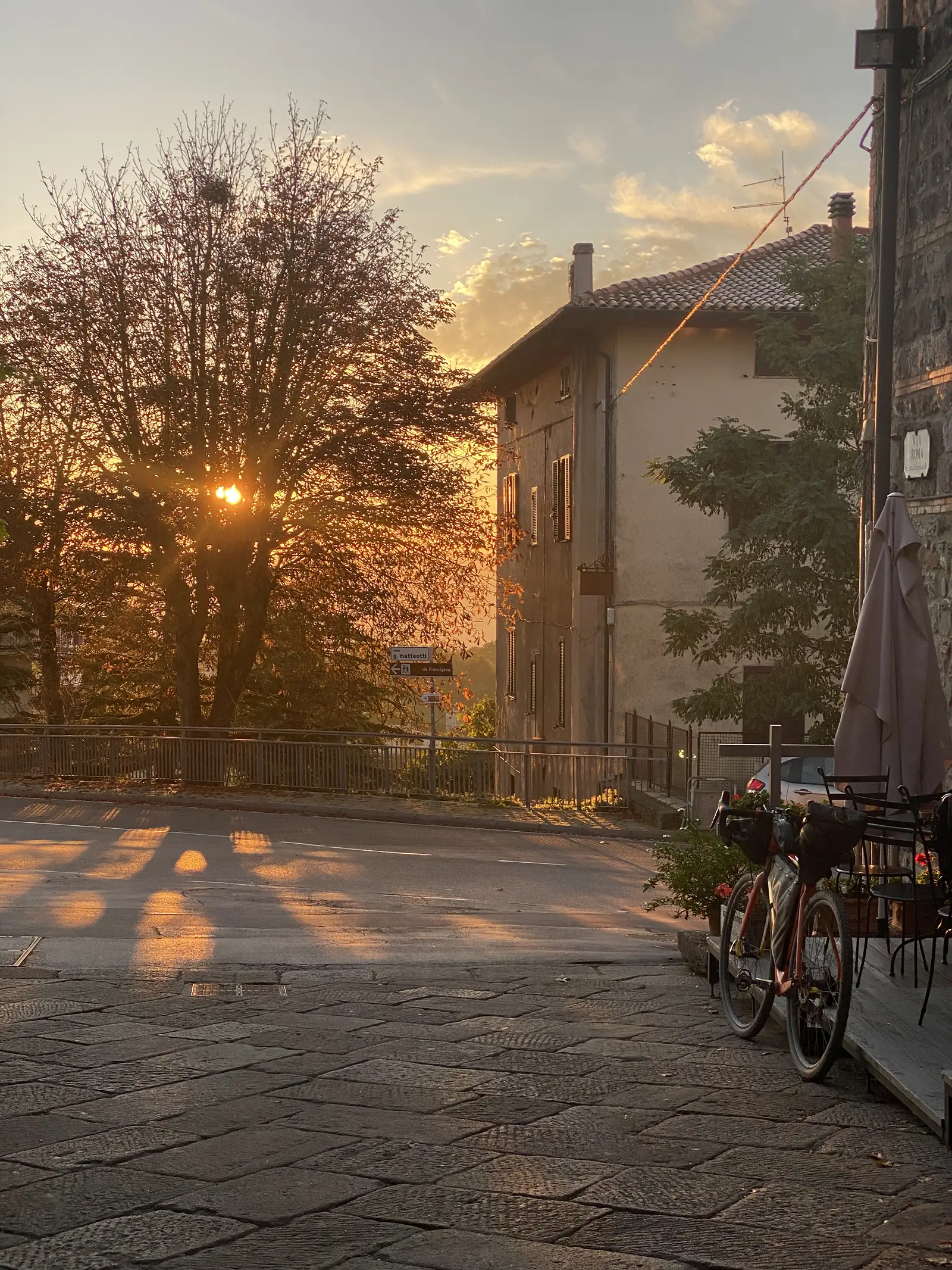 Low evening sun shining through a bare tree in a cobbled piazza, with a bicycle parked beside café tables under a parasol.