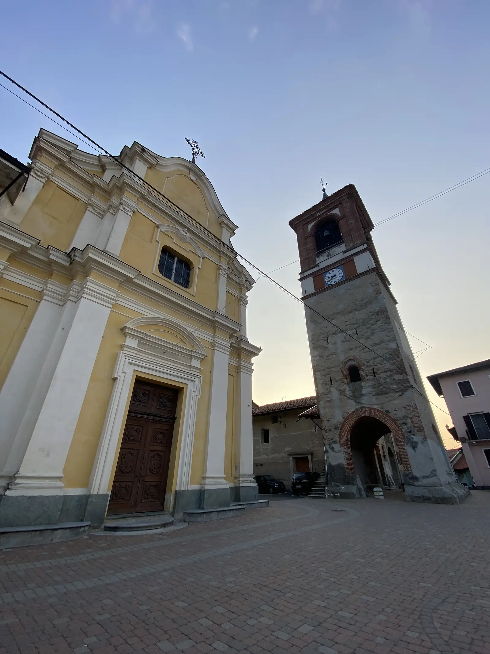 A yellow baroque church facade with a stone cross on its pediment, seen from below alongside a tall brick bell tower with a clock.