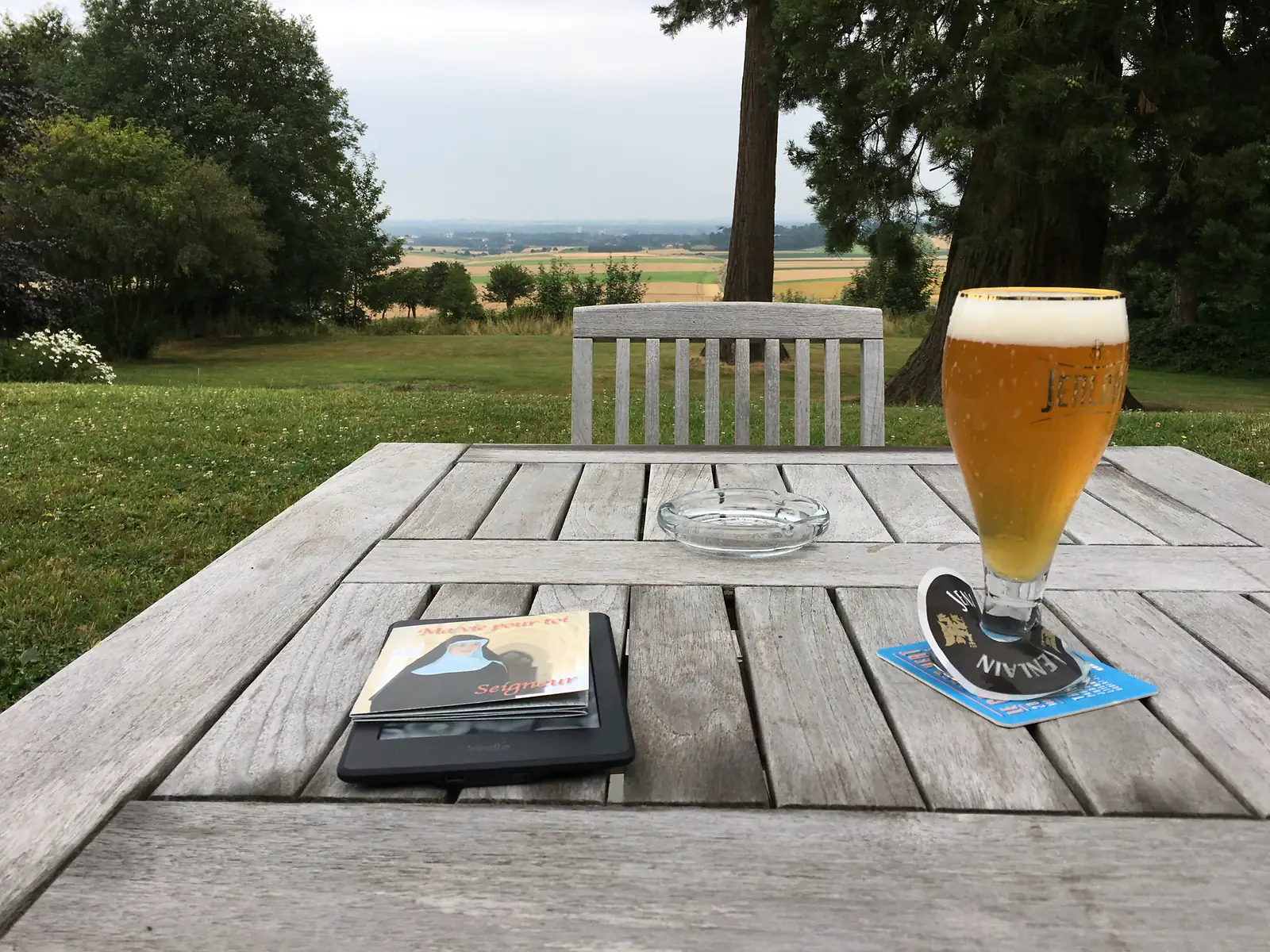 A half-drunk glass of pale beer and a Kindle e-reader sit on a weathered wooden garden table, with an empty chair facing a view of rolling farmland beyond.