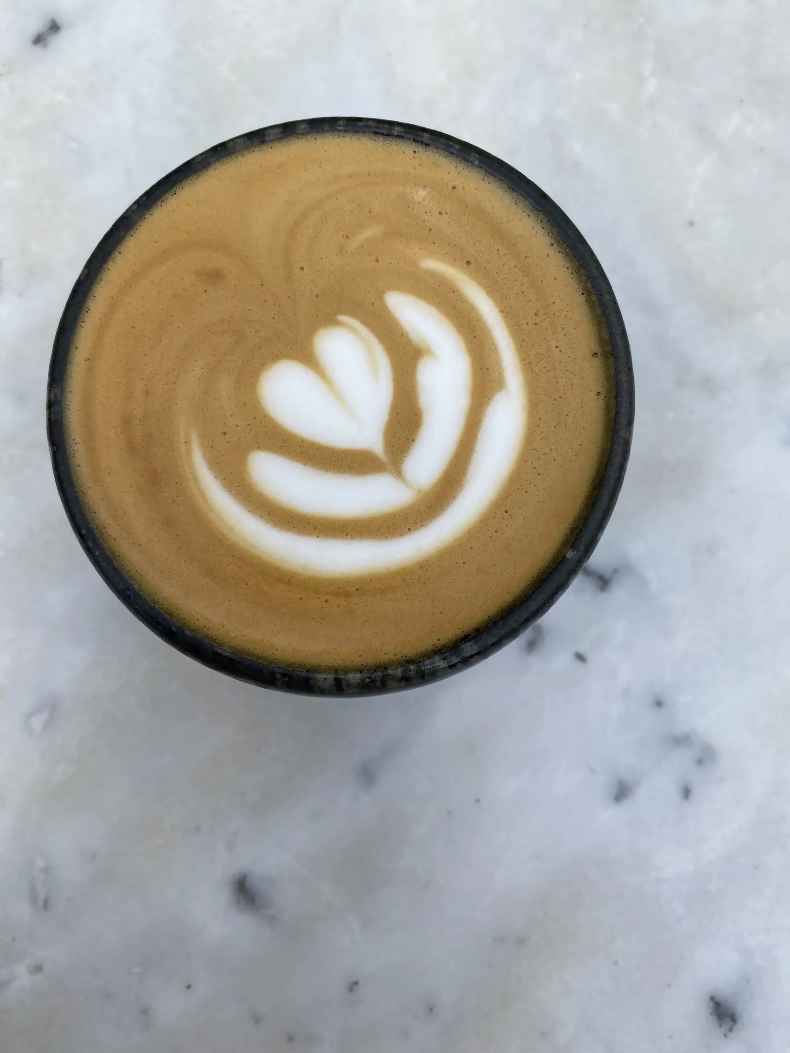 A flat white in a dark ceramic cup with a leaf pattern drawn in the crema, seen from above on a white marble table.