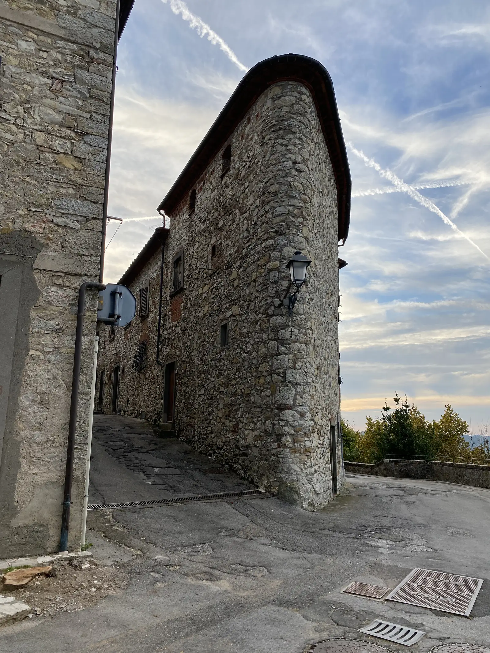 A rounded stone corner tower of an old building rising above a narrow paved lane, seen from below against a sky streaked with vapour trails.