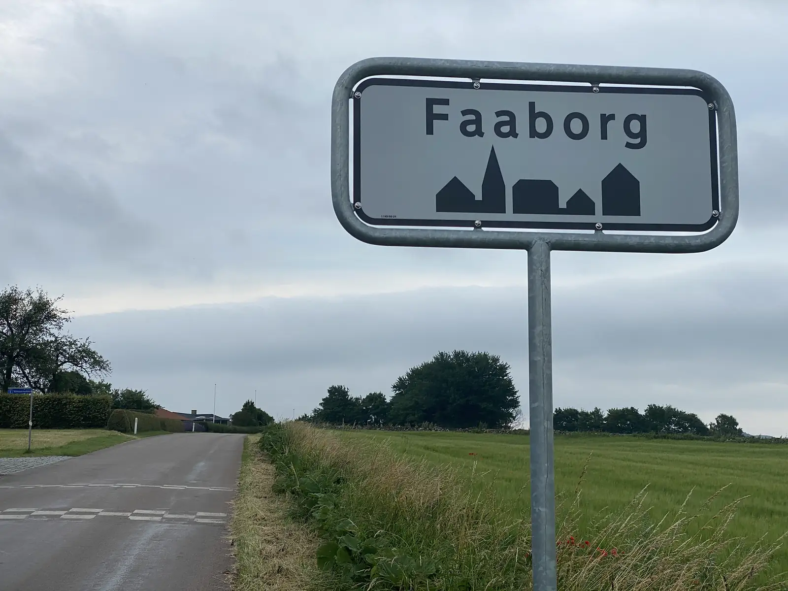 A town boundary sign reading "Faaborg" with a village silhouette, beside a quiet road and green cereal field.