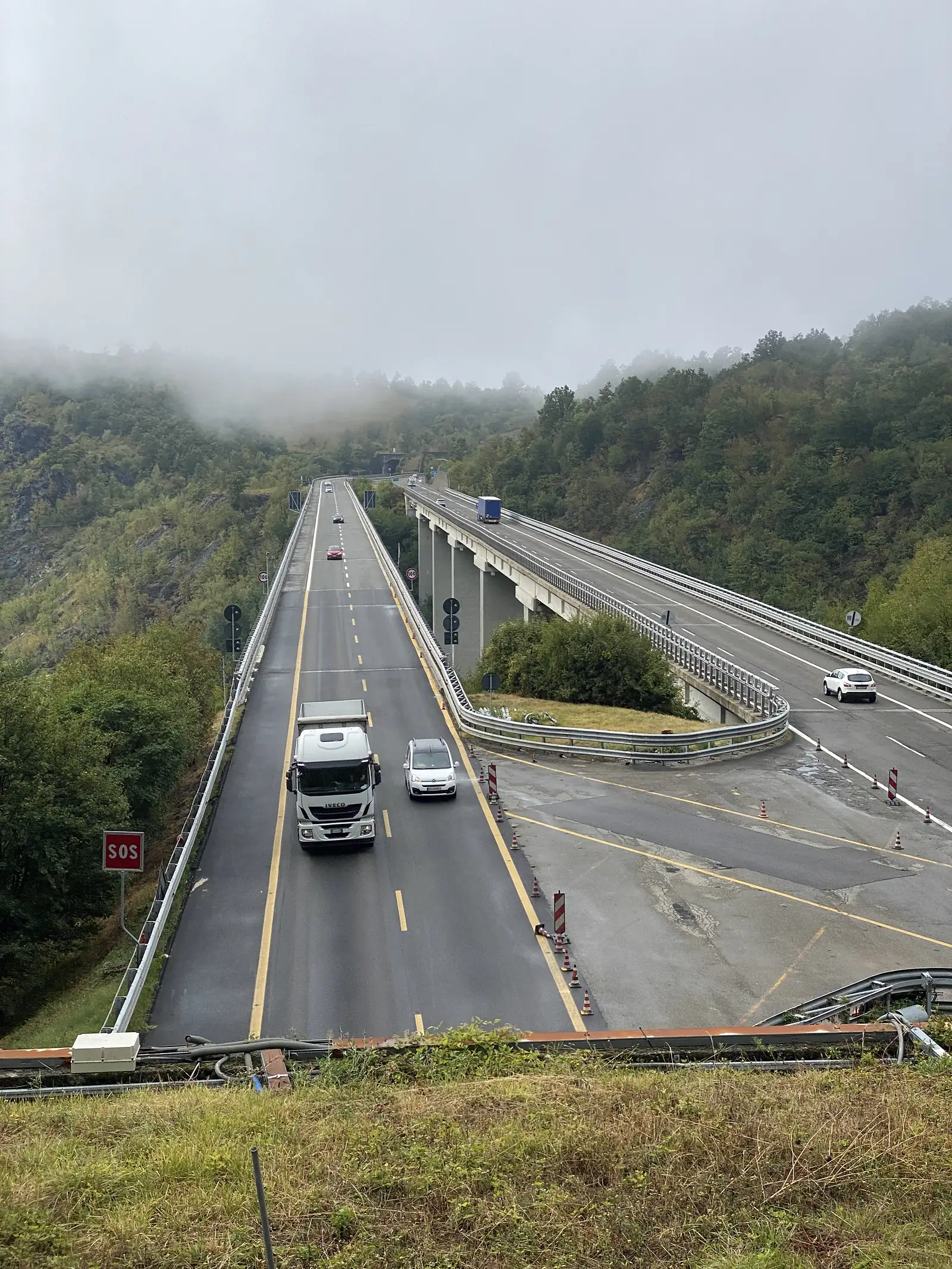 Cars and a lorry cross a curving motorway viaduct above forested hillsides, with roadworks cones marking a closed lane.