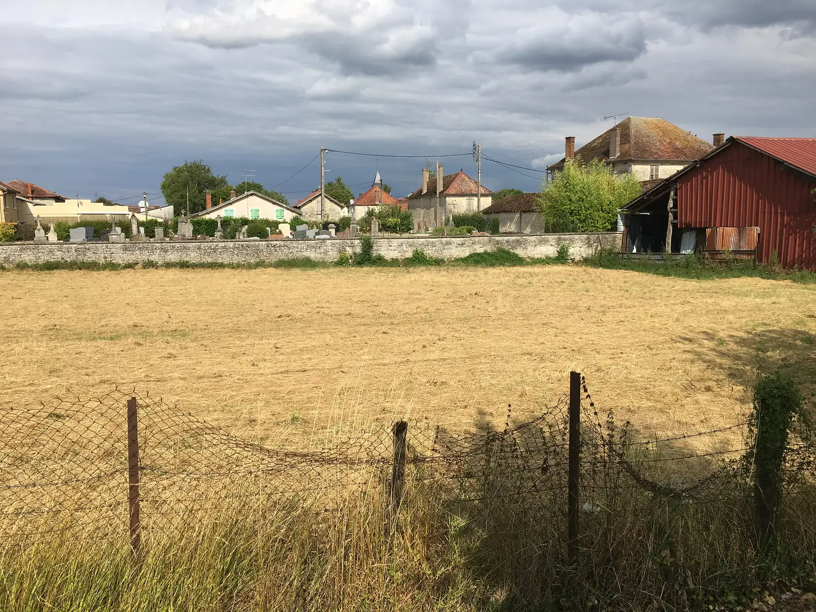 A walled cemetery and cluster of stone houses with a church spire are seen across a stubble field under a stormy sky.