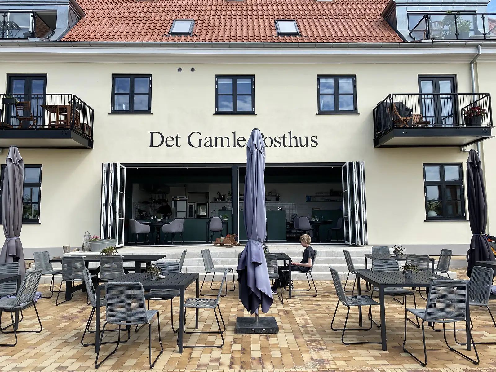 A cream-rendered building lettered "Det Gamle Posthus" with folding doors open onto a brick terrace of cafe tables, a child sitting on the steps.