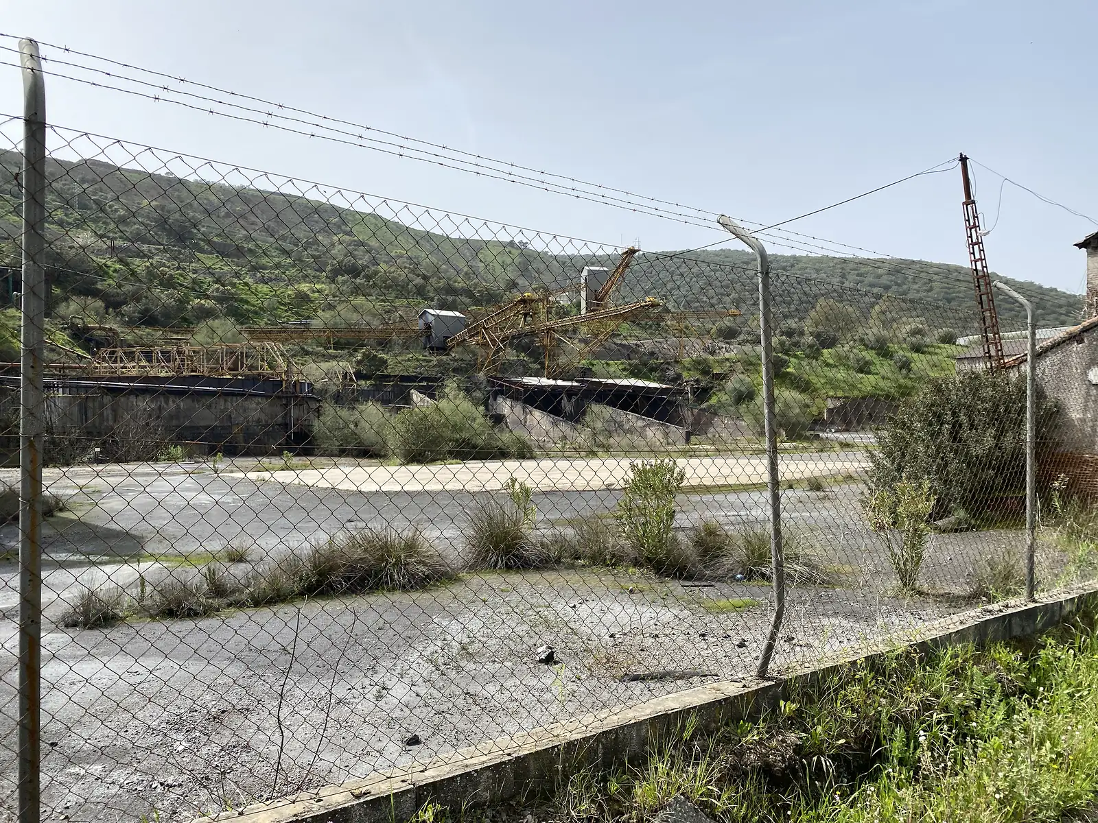 Rusting yellow gantry cranes and conveyors of a derelict mine works seen through a chain-link and barbed-wire fence, with green hills behind.