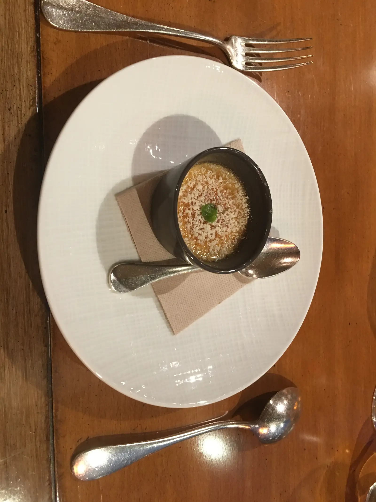 A small crème brûlée topped with grated cheese and a mint leaf, served in a dark ramekin on a white plate.