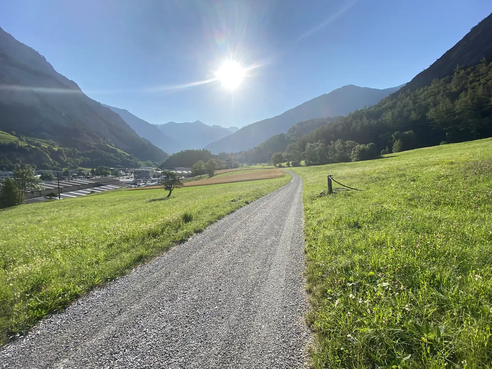 A gravel farm track descending through a sunlit valley towards distant village rooftops, with a snow-streaked mountain peak on the horizon.