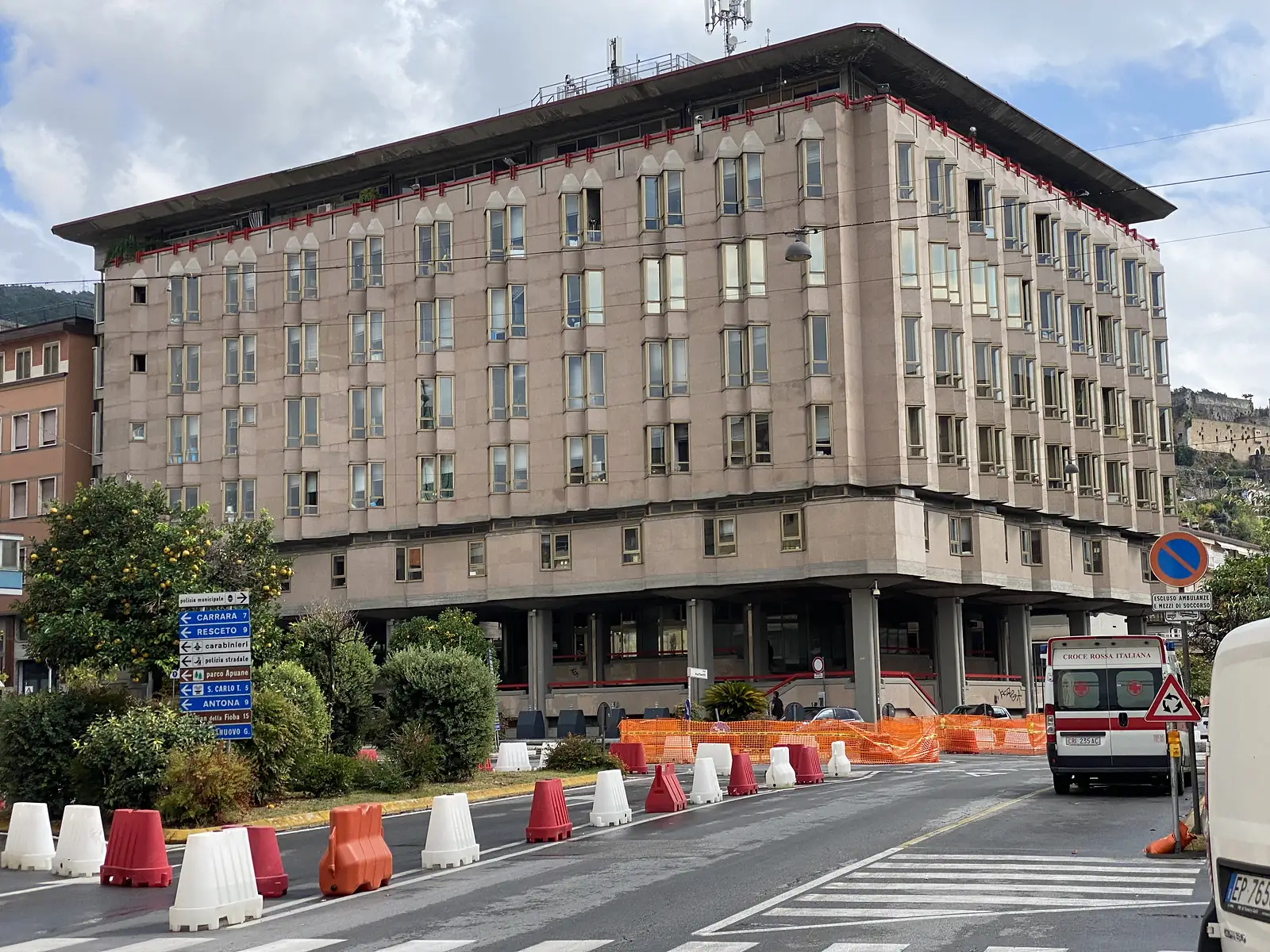A tiered concrete office building on stilts, its road signs pointing to Carrara, Resceto and the Apuane park among other local destinations, with an ambulance parked outside and quarried hills visible behind.