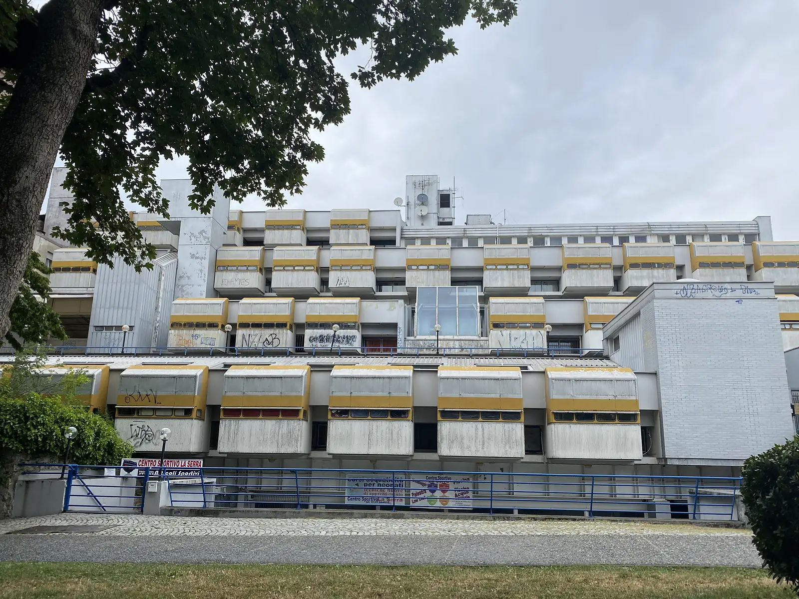 The rear of a mid-century sports-centre building, tiers of yellow-banded concrete balcony pods covered in graffiti beneath an overcast sky, a tree in the foreground.