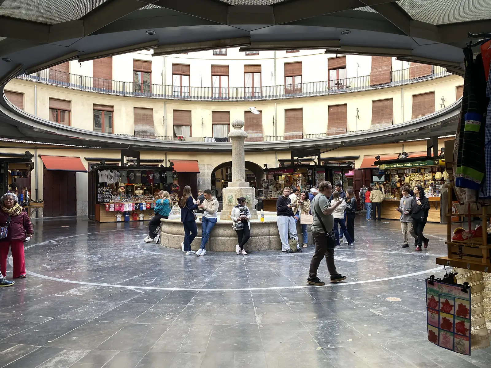 A circular market square ringed by shuttered apartment fronts under a canopy, with stalls and people gathered around a stone fountain.