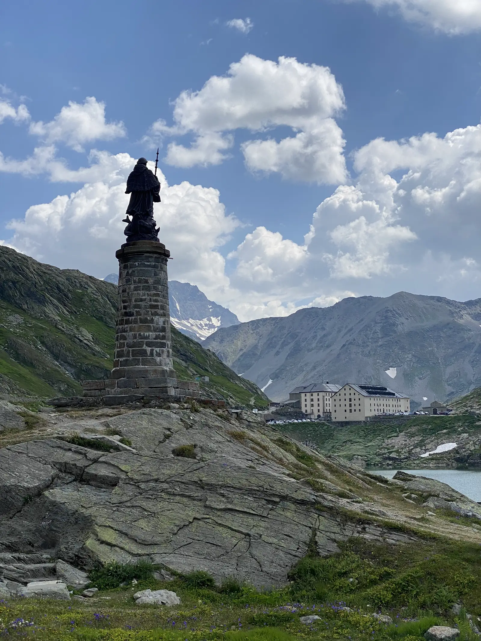 
A dark bronze statue of a robed, staff-carrying figure atop a stone plinth, looking out over the pass lake and hospice below.
