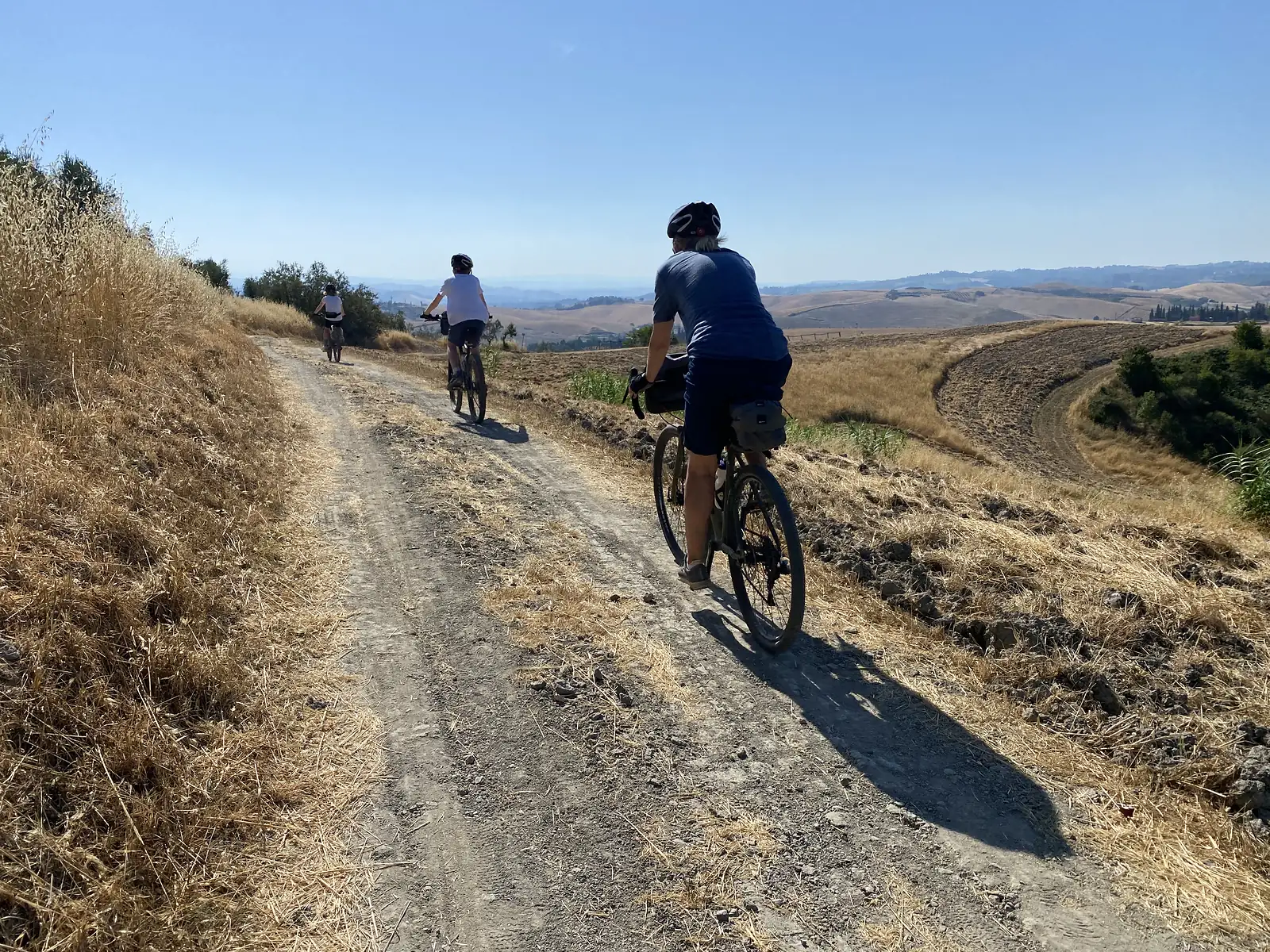 Three cyclists ride single file along a dirt track cresting a hill, with sweeping golden farmland and rolling hills stretching to a hazy horizon.