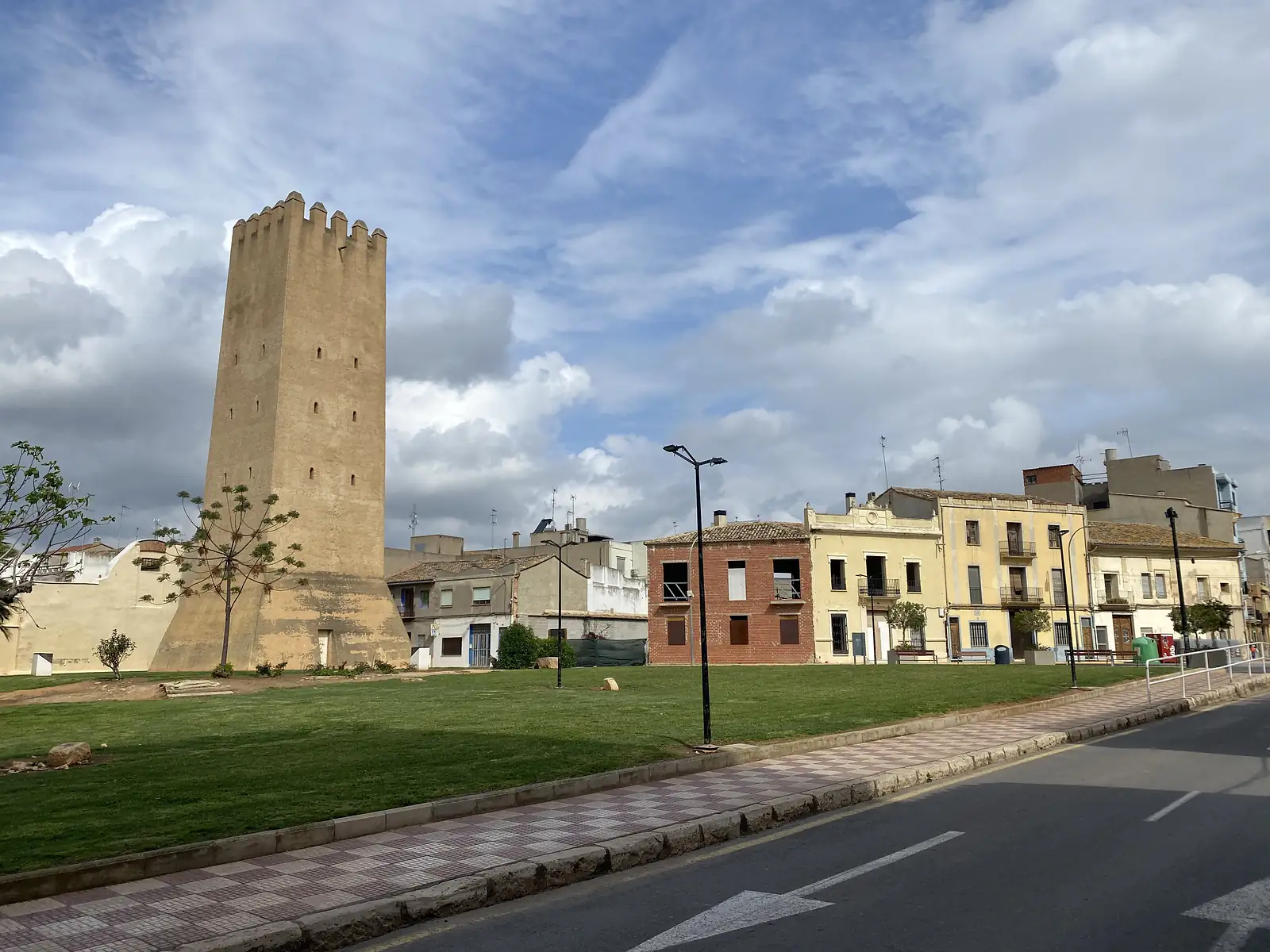 A tall battlemented brick tower standing alone on a lawn beside a road, with a row of low town houses behind.