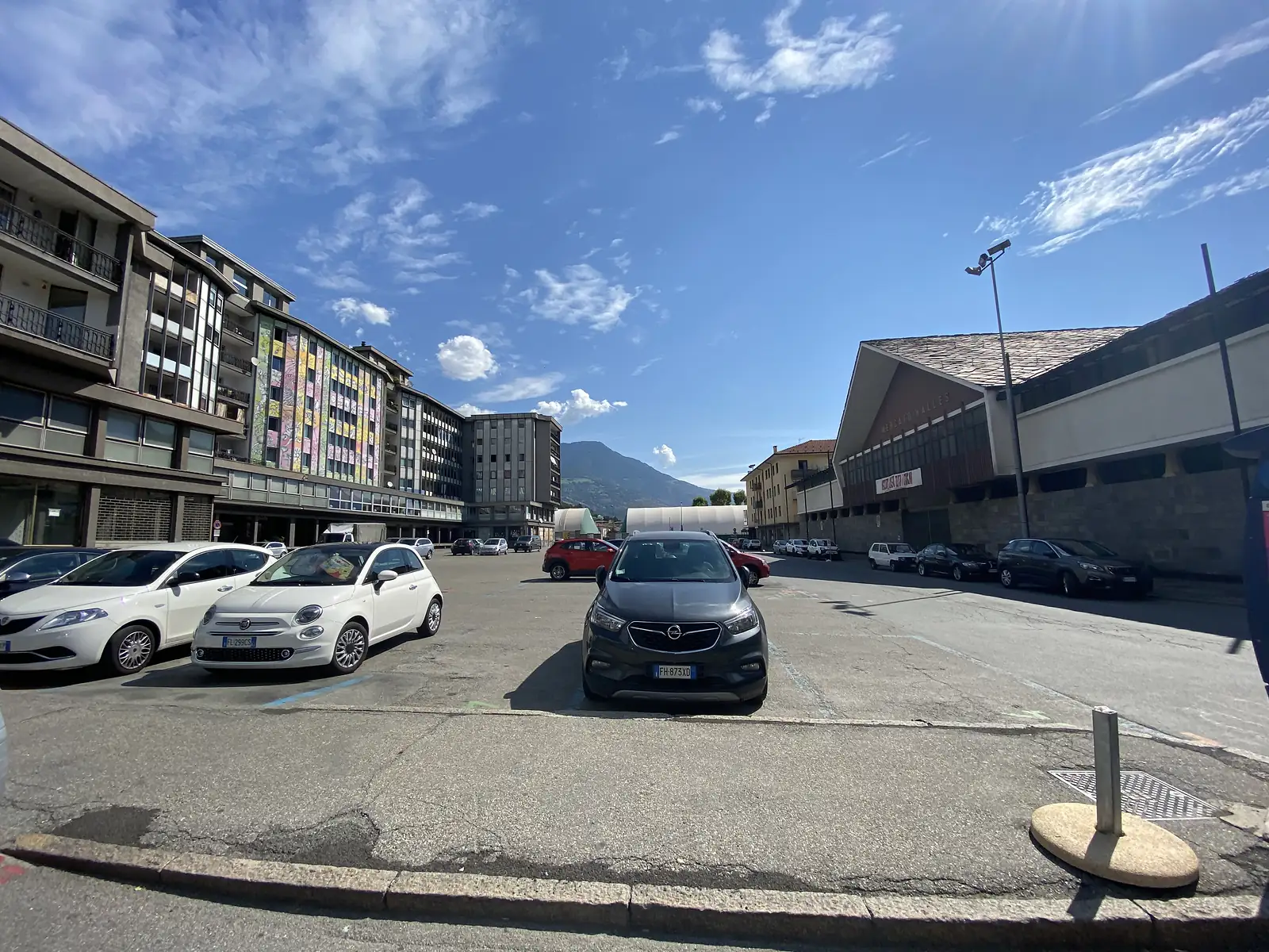 Cars parked in a town square beneath a colourful mural-painted apartment block, a market hall and forested mountain visible beyond.
