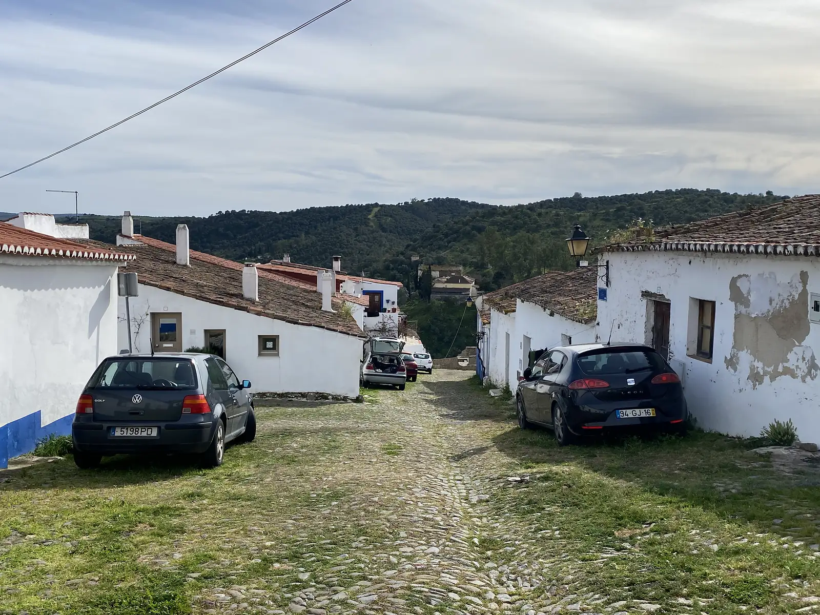Cars parked along a grassed-over cobbled street between low whitewashed houses with pantile roofs, looking downhill towards wooded slopes.