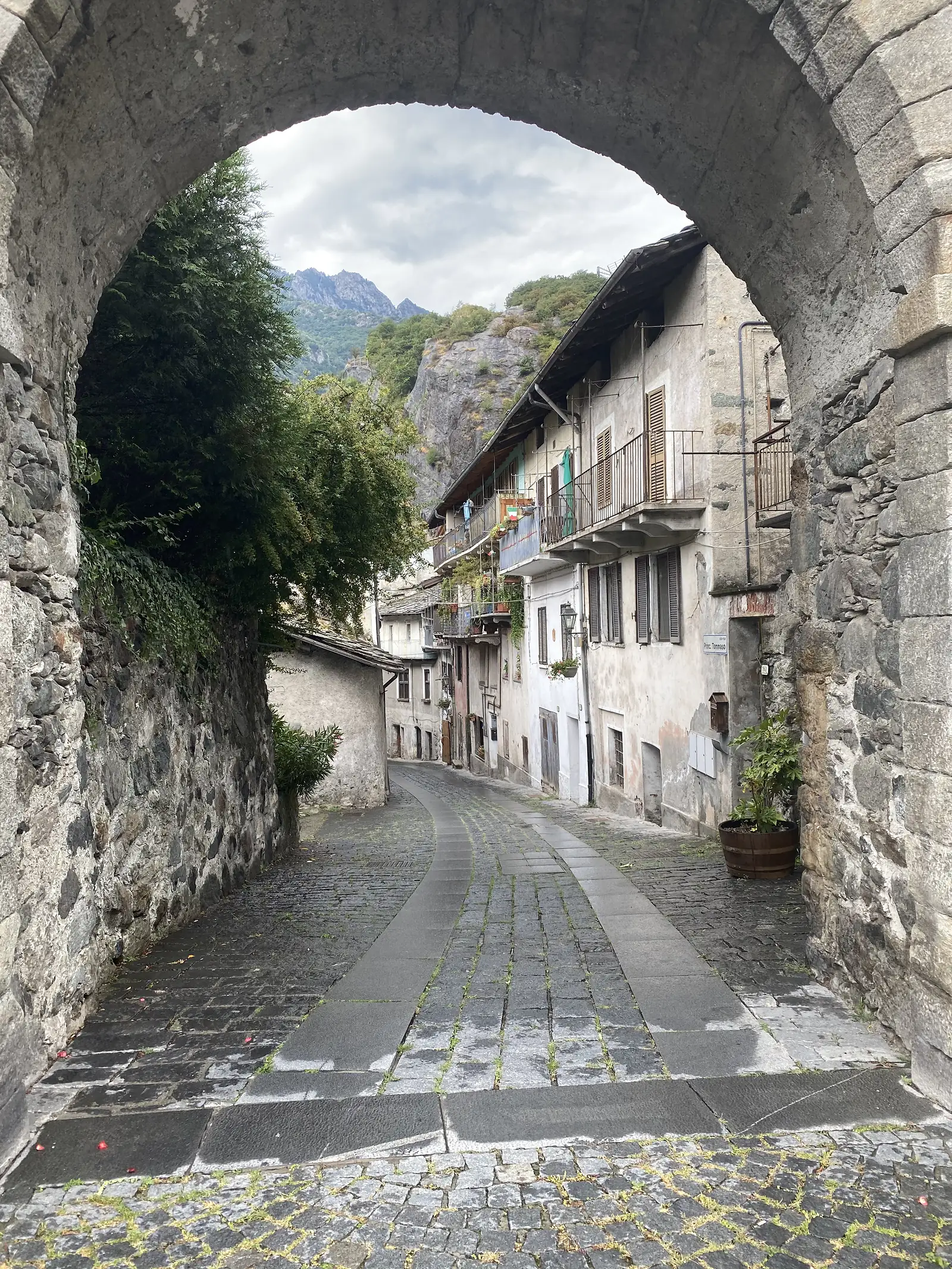 A cobbled village street framed by a stone archway, lined with shuttered houses and balconies, with a rocky peak rising behind the rooftops.