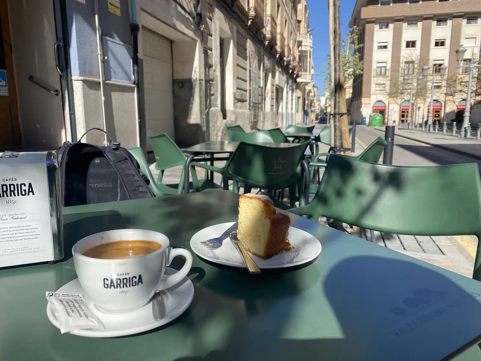 A coffee and a slice of lemon sponge on a green pavement cafe table, a rucksack on the chair behind and a city street beyond.