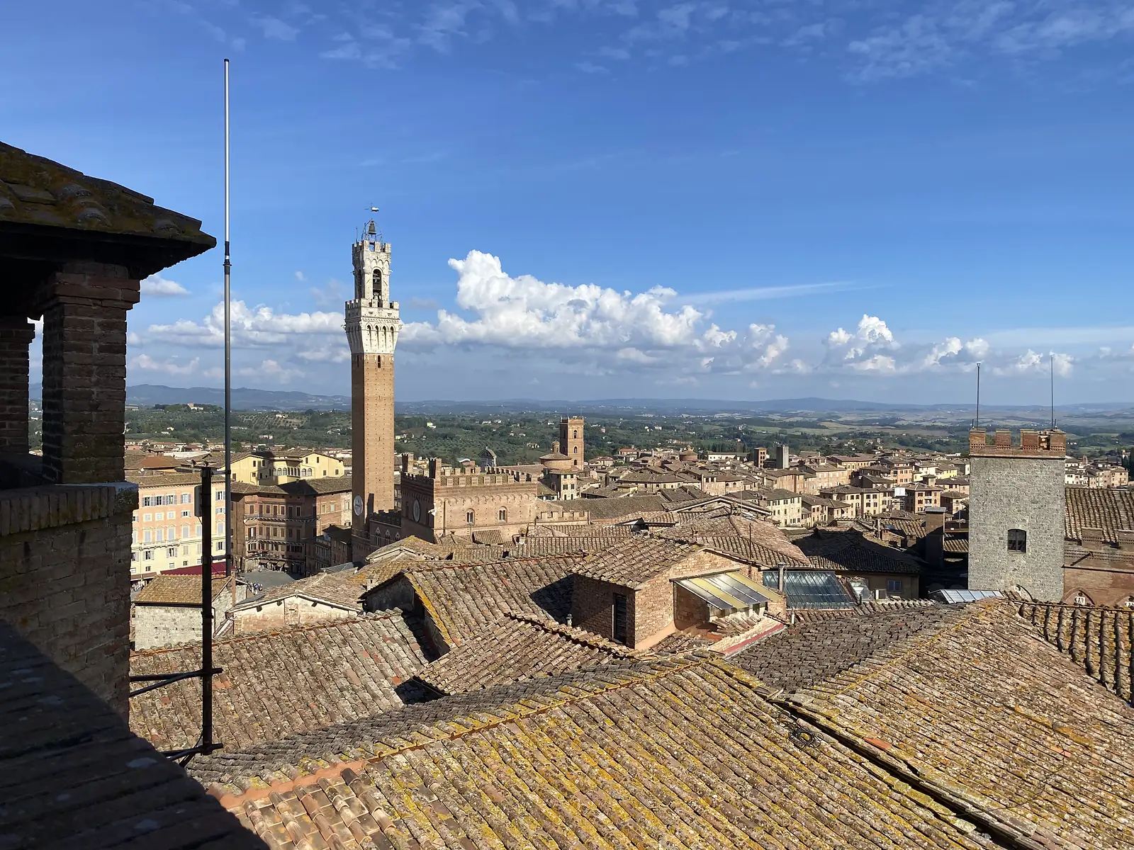 Rooftop view over terracotta tiled roofs to a tall brick bell tower with a crenellated crown, rising above the historic centre of a hill town.