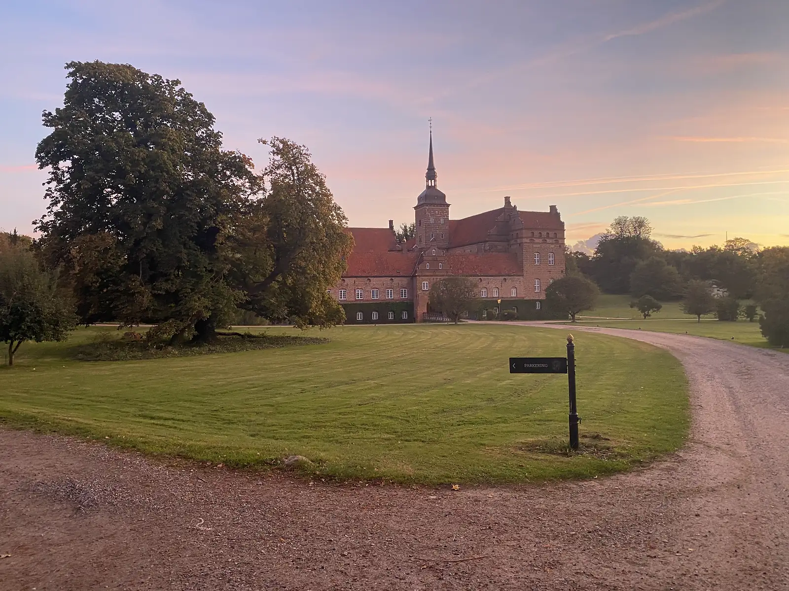 A red-brick manor house with a spired tower and red-tiled roofs standing across a mown lawn in low evening sun.