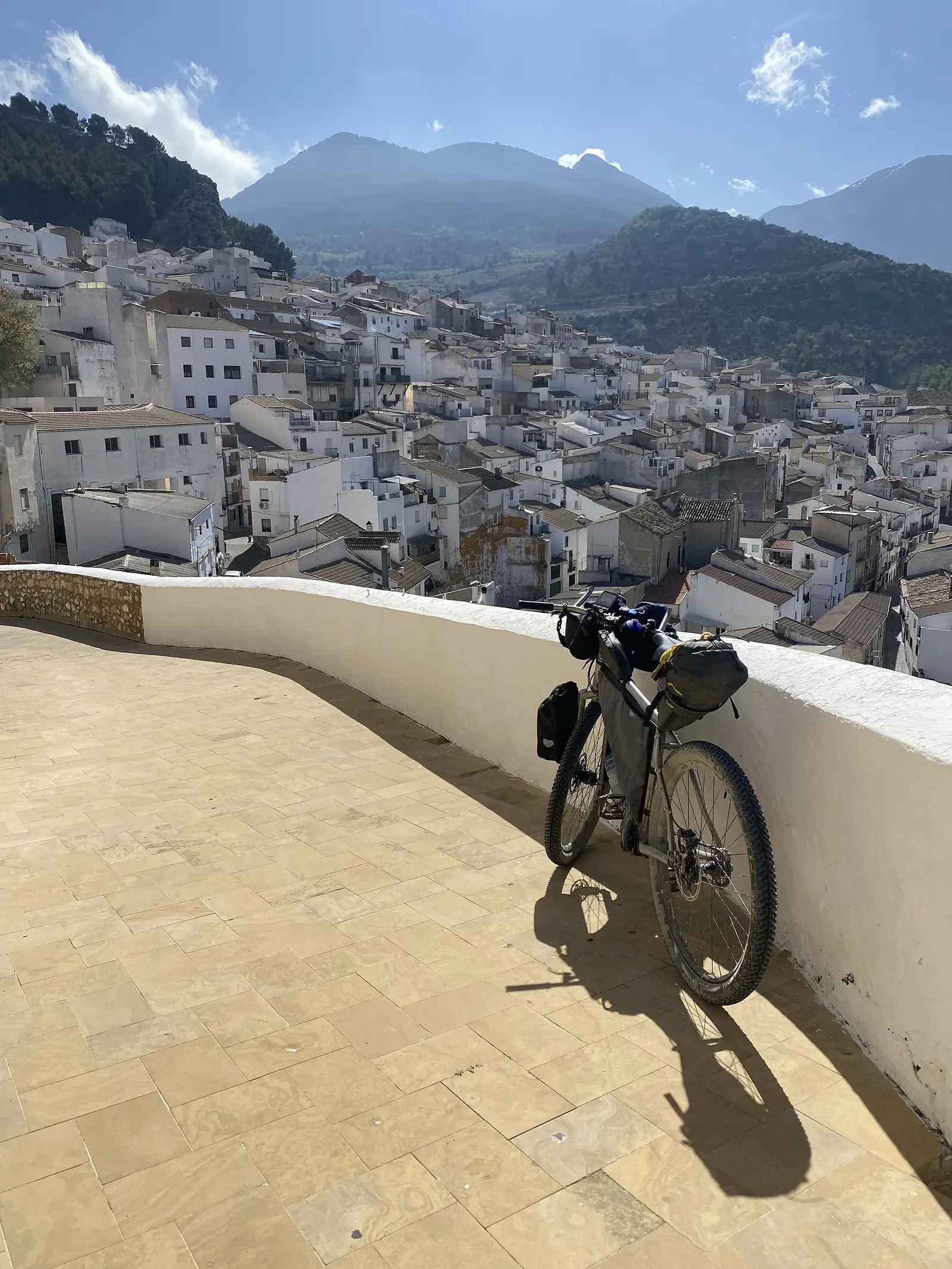 A loaded titanium adventure bike leaning on a whitewashed parapet above a hill town of white houses packed down the slope, with wooded mountains behind.