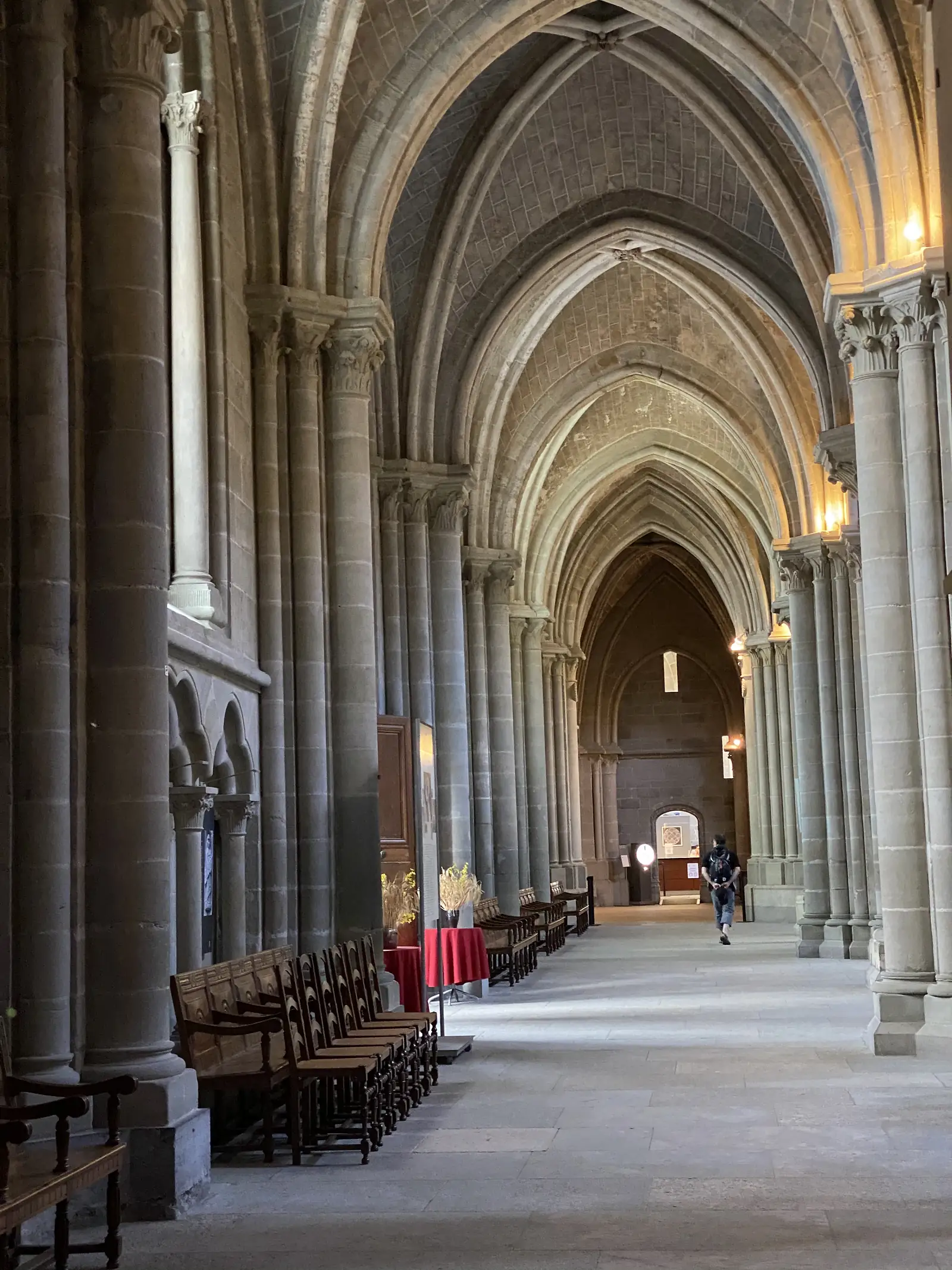 A vaulted side aisle of Lausanne Cathedral lined with wooden chairs, a lone visitor walking away down the stone passage.