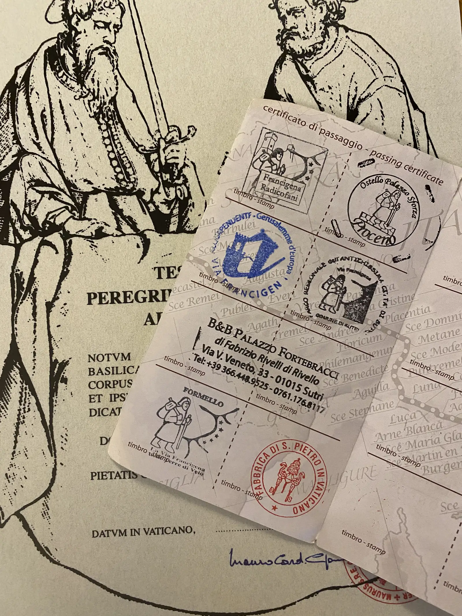 A pilgrim's credential document covered in stamps from stops along the Via Francigena, including Sutri, Formello and "Fabbrica di S. Pietro in Vaticano", laid over a printed Latin testimonium certificate.