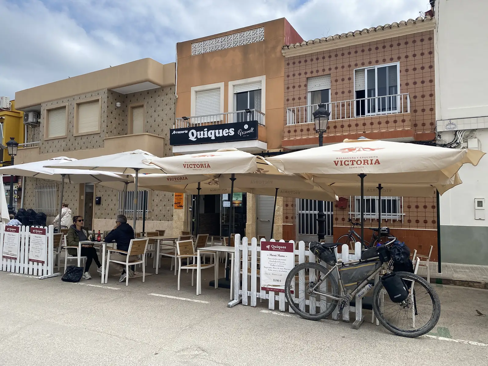 A loaded bikepacking bike propped against a white picket fence outside a restaurant terrace of parasols and diners, its sign reading "Arrosseria Quiques".