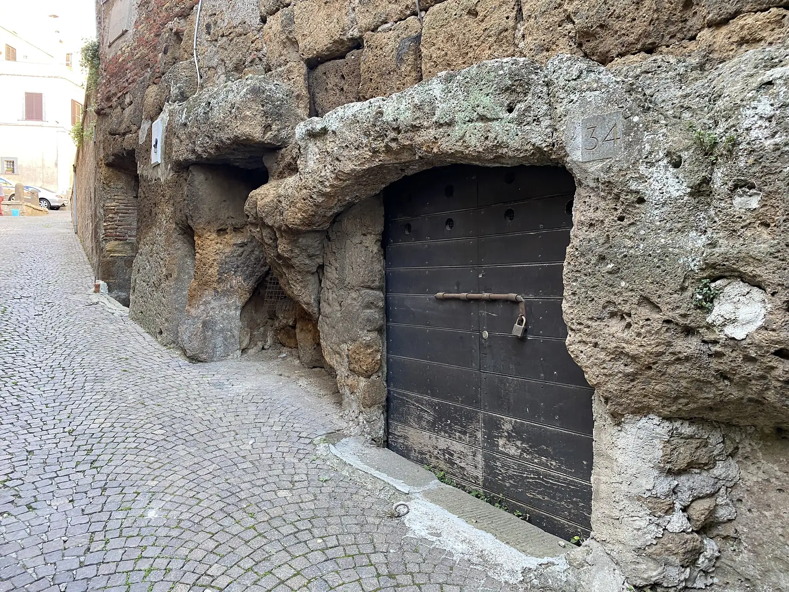 A padlocked wooden door numbered "34" set into a rough-hewn stone archway carved directly from the rock face, opening onto a cobbled lane.
