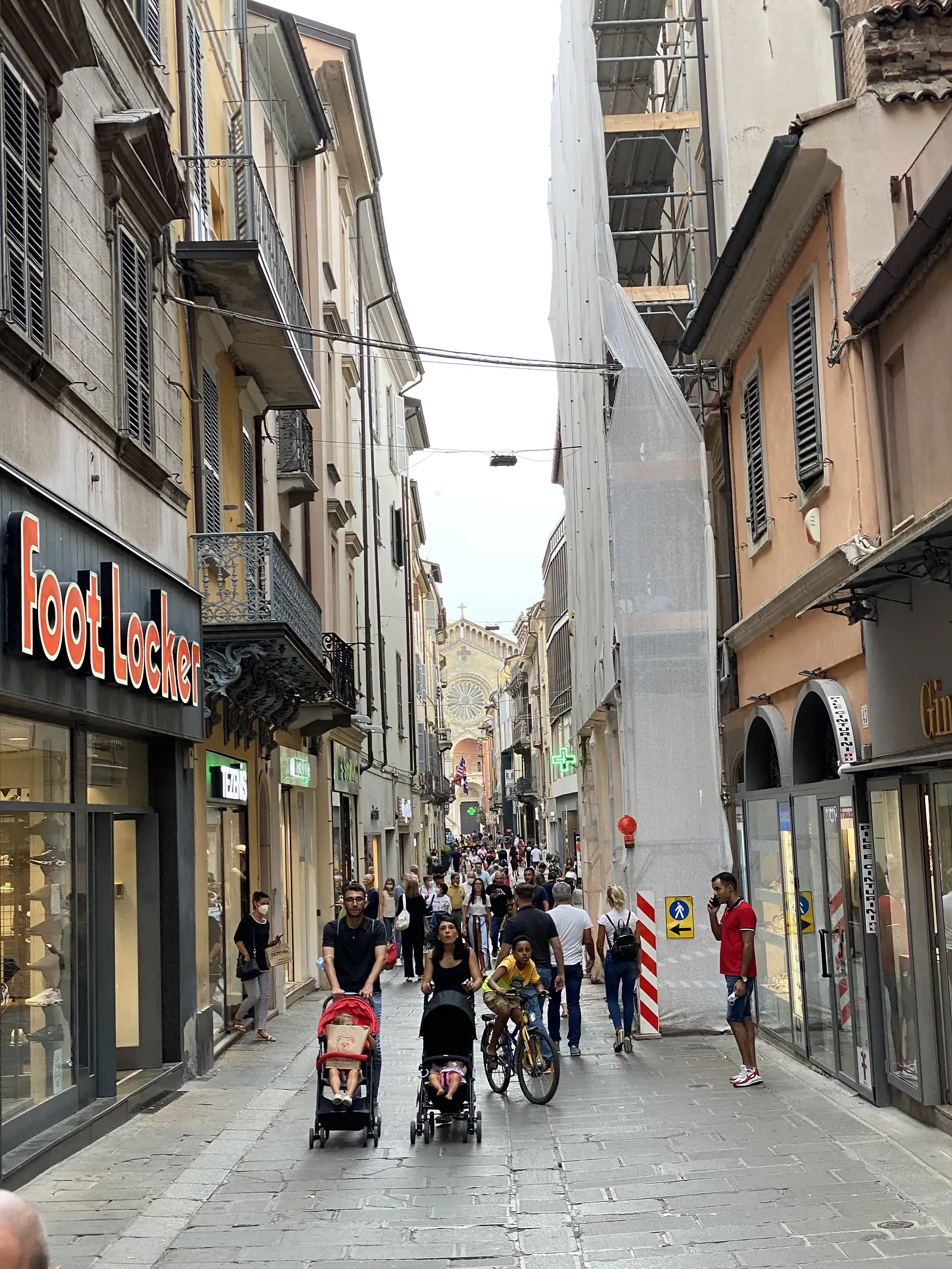 Shoppers and pedestrians fill a narrow street in Piacenza past a Foot Locker store, with a church's rose window visible in the distance.