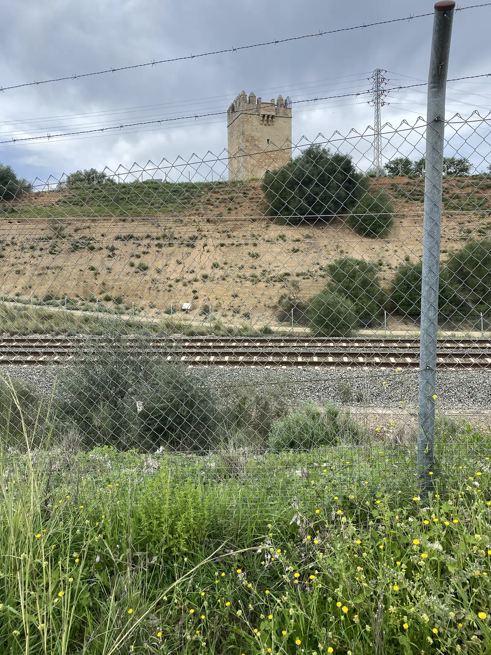 A battlemented stone tower on an olive-covered hillside seen through chain-link and barbed wire, with railway tracks running below.