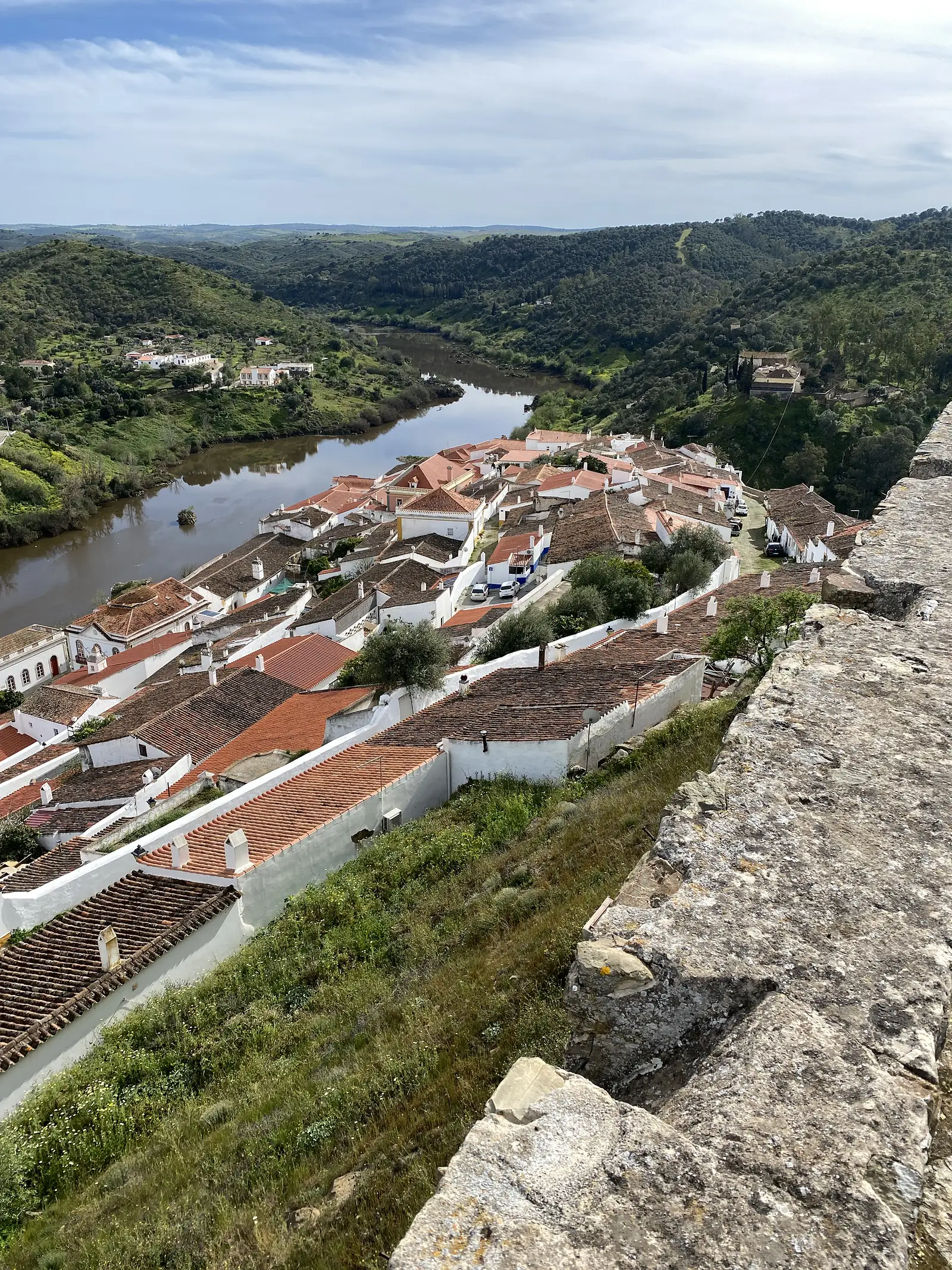 Terracotta rooftops of a whitewashed village packed tightly below a rocky crag, with a wide brown river curving past and wooded hills beyond.