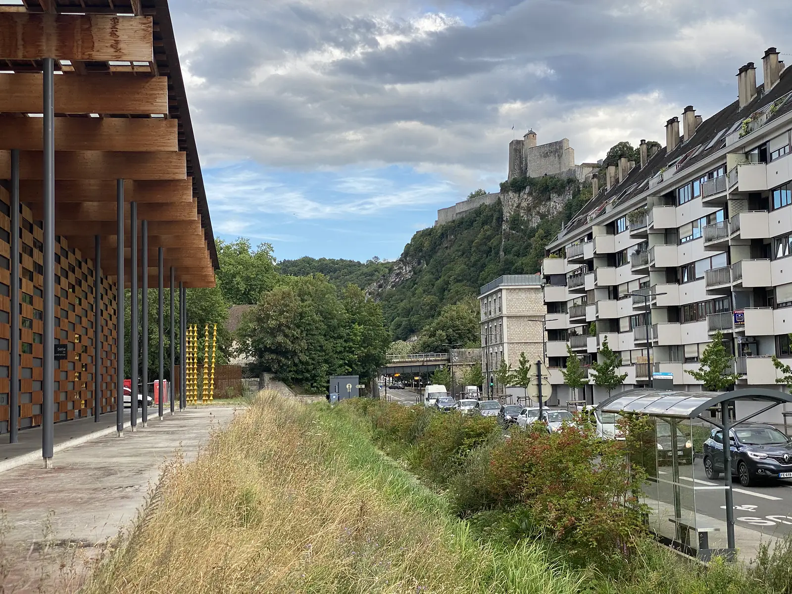 Besançon's hilltop citadel, with turrets and a flag, rises above a cliff behind a row of modern apartment blocks and a timber-clad building.