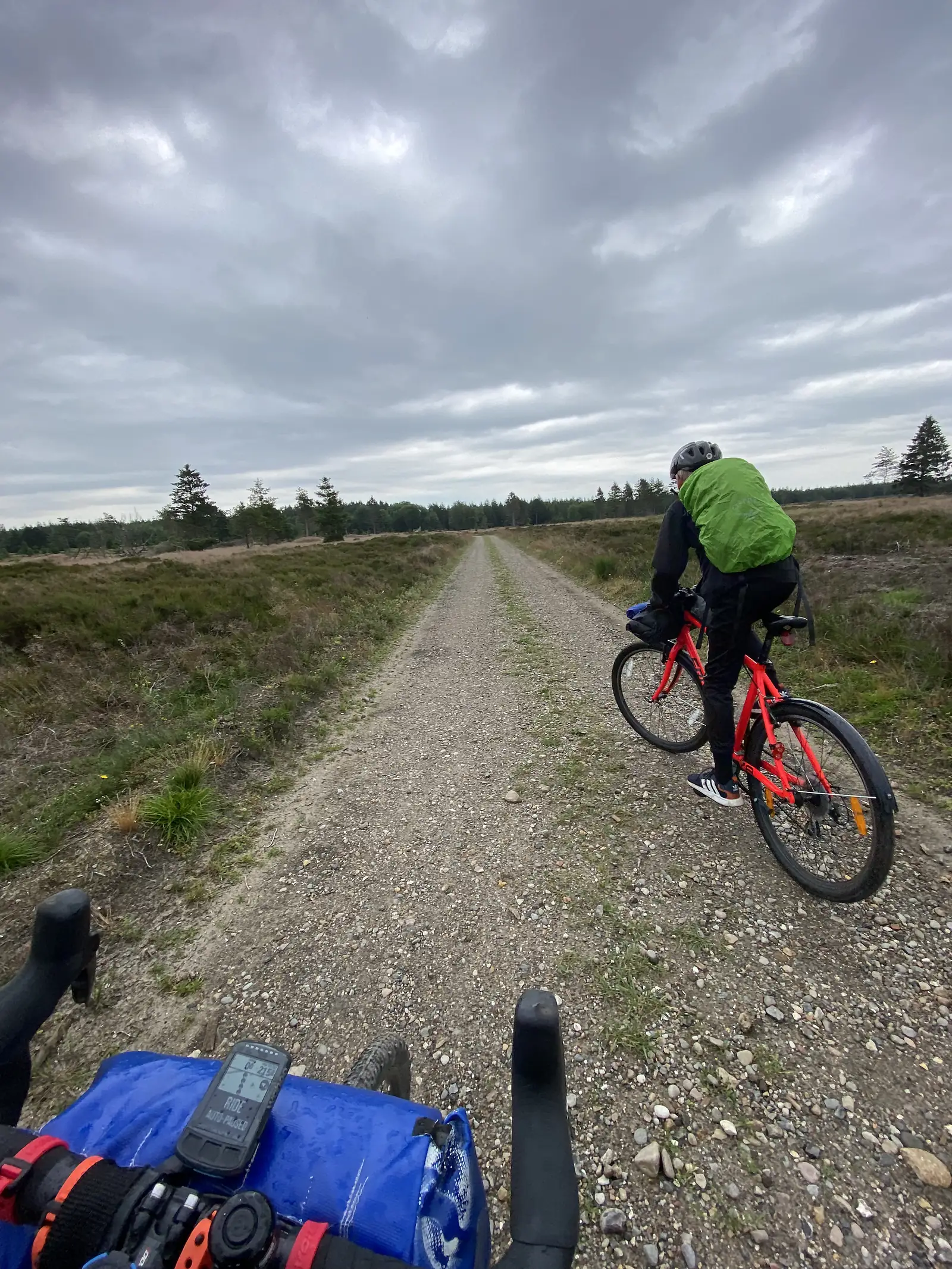 A handlebar view of a rider in a green jacket on a loaded red bike ahead on a gravel track across open heath under grey cloud.