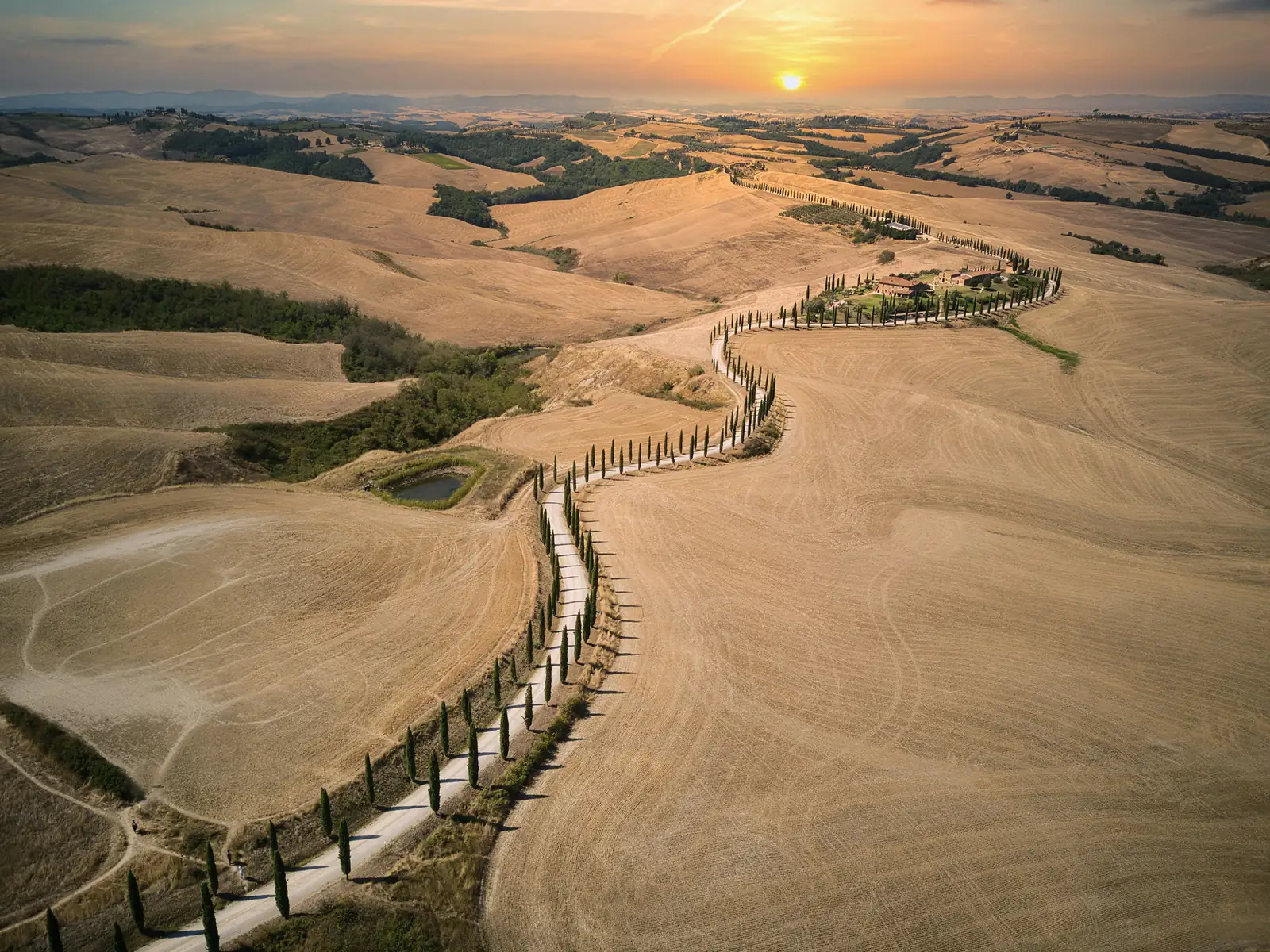 A long distance shot from a drone of a gravel road lined by trees in Tuscany. At sunset.