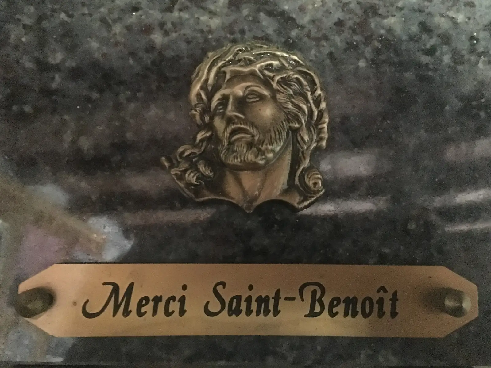 A bronze plaque bearing a relief of the crowned head of Christ is inscribed 'Merci Saint-Benoît' on a dark marble memorial.
