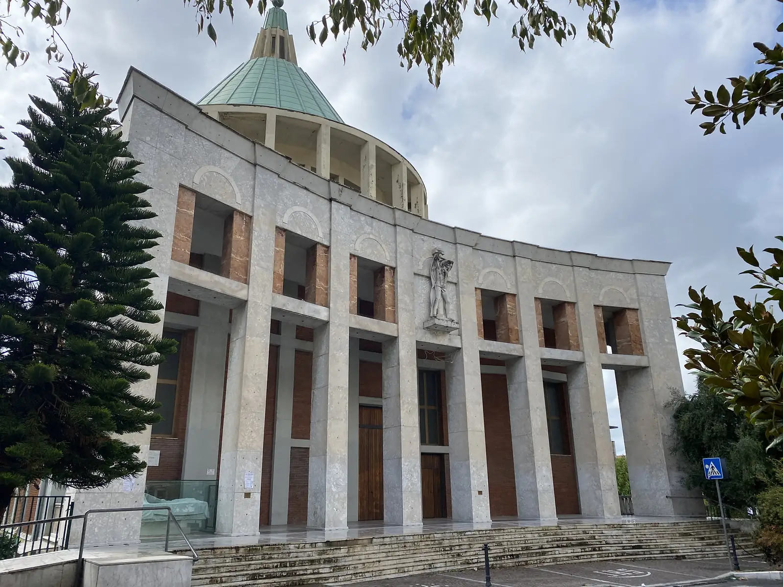 A modern marble-clad church with a tall green copper dome and a stone statue of a saint set into its arcaded façade, fronted by a broad flight of steps.
