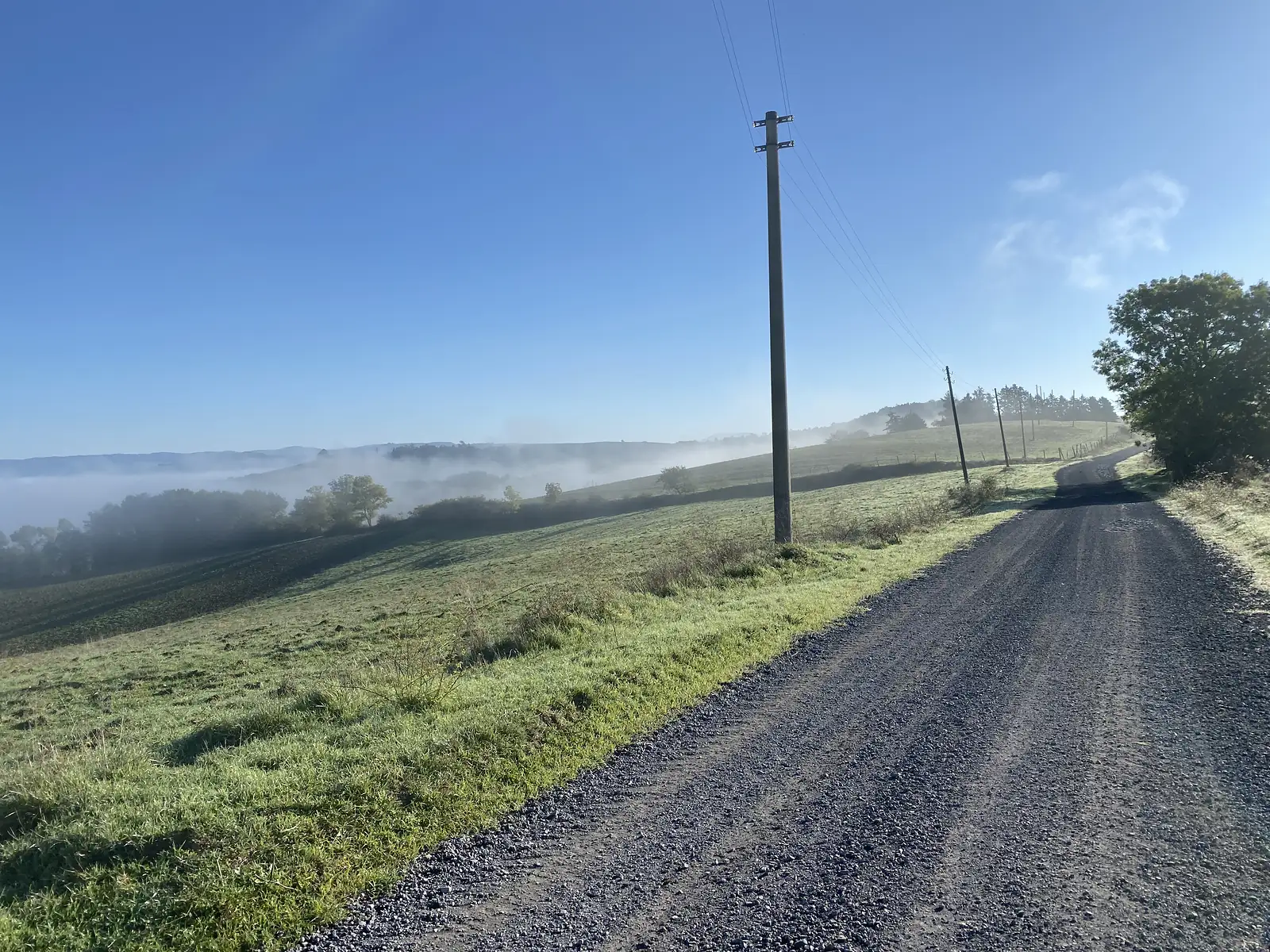 A gravel track runs along a hillside above a valley filled with morning mist under a clear blue sky.