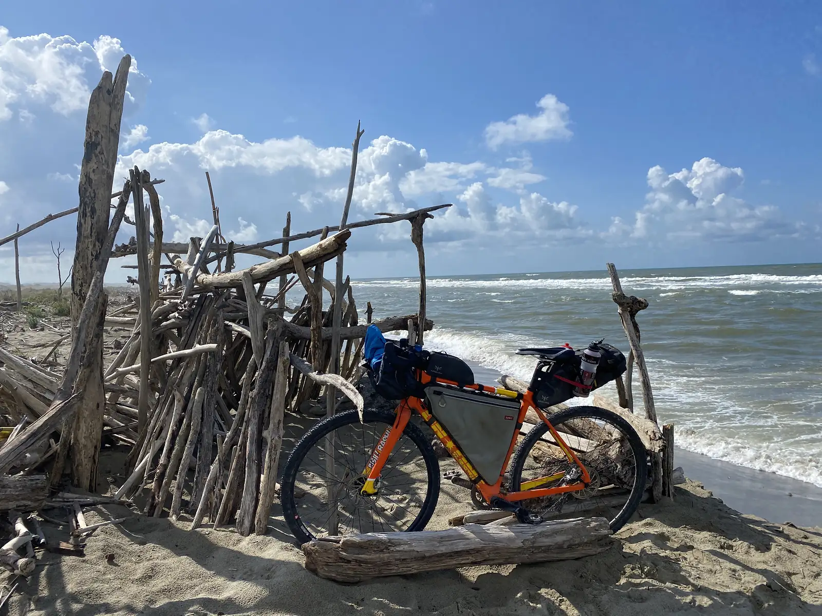 A loaded orange bikepacking bike propped against a driftwood shelter built on the beach, waves breaking behind it.