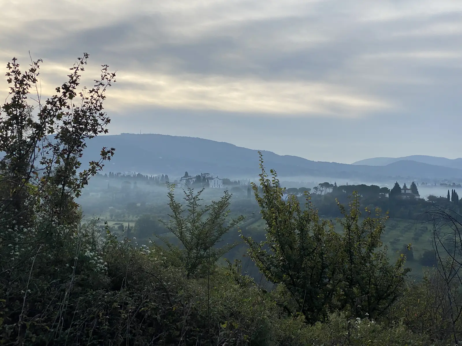 A misty Tuscan valley at dawn with a white villa and cypress trees emerging above low fog, hills silhouetted beyond.