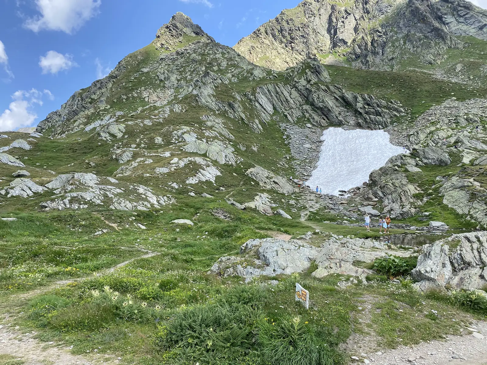 
Walkers pausing beside a lingering snow patch on a rocky slope, next to a yellow waymark post numbered 103.
