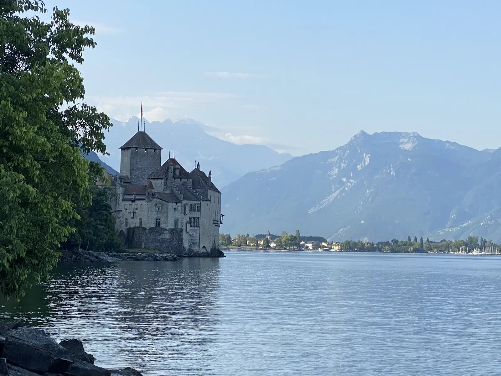 Framed by an overhanging tree, a turreted stone castle stands on a rocky lakeside point with snow-streaked peaks rising behind it.