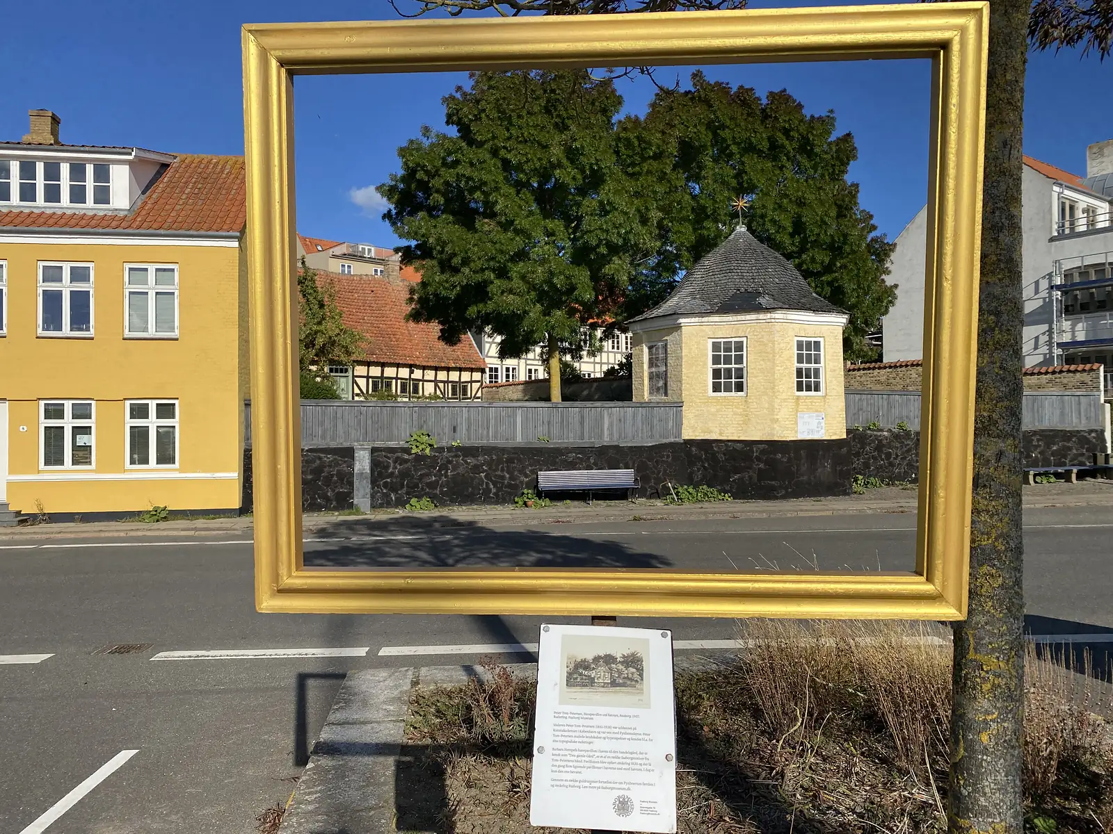 An empty gilt picture frame mounted outdoors so that it frames an octagonal yellow pavilion across the road, with an interpretation panel below.