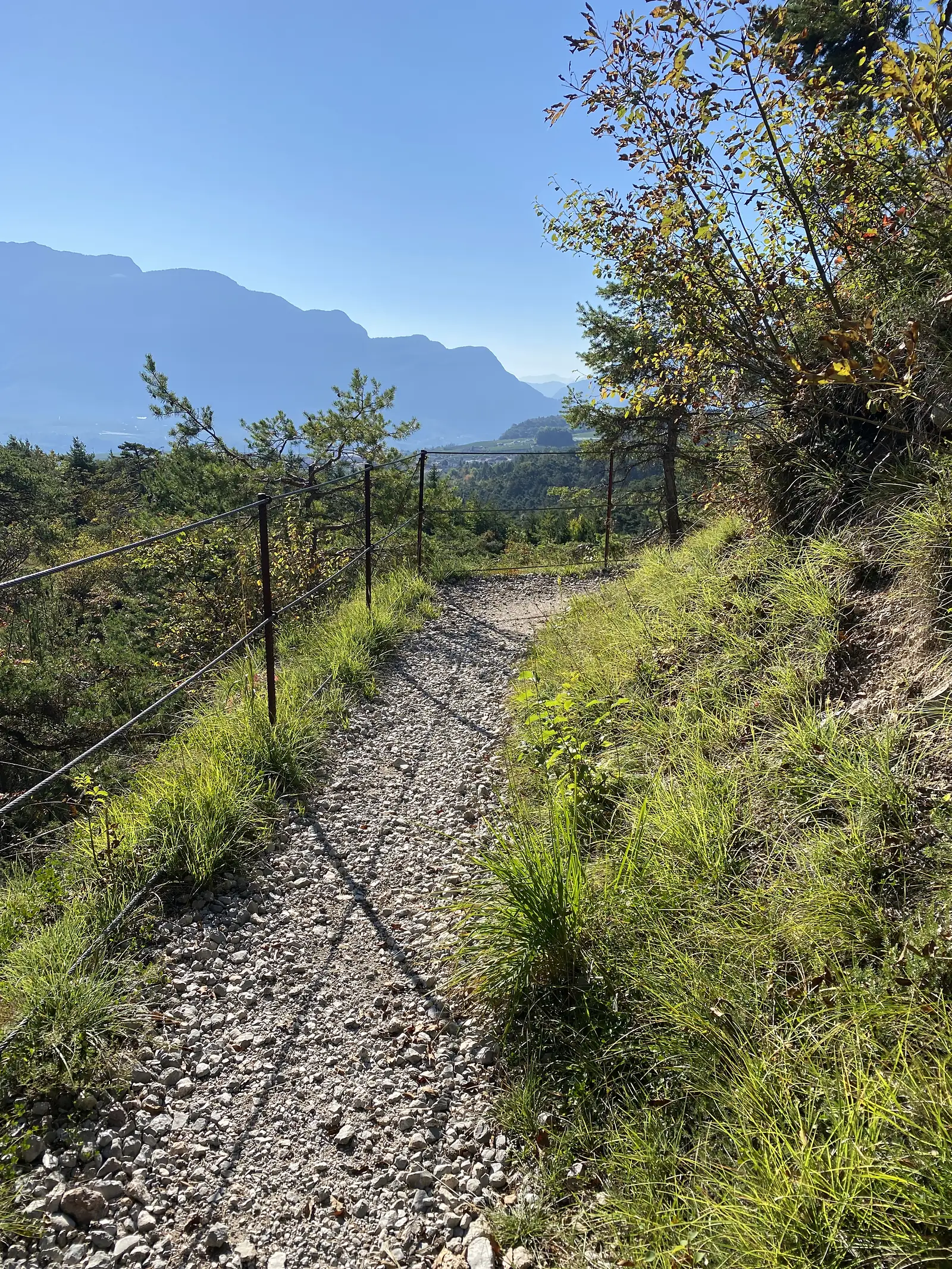 A stony singletrack climbs beside a rusted wire fence, opening out to a hazy valley and a long mountain wall in the distance.