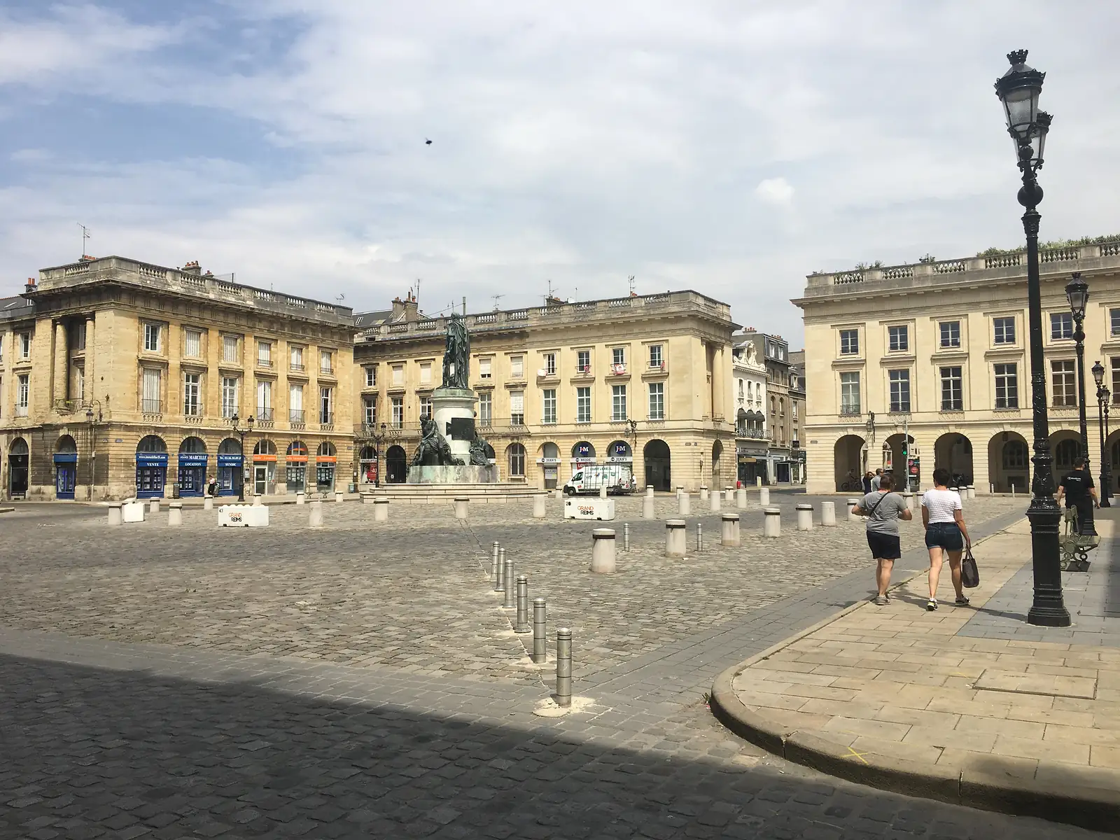 Pedestrians cross a cobbled square in Reims lined with grand stone buildings, a bronze statue on a plinth, and a barrier reading GRAND REIMS.