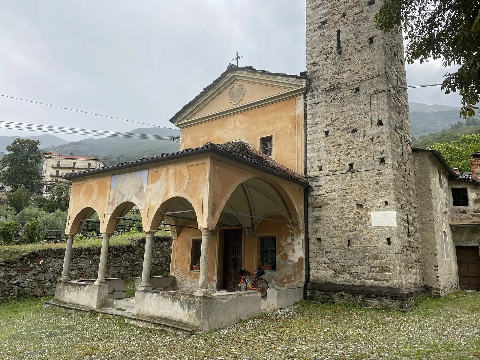 A small ochre church with a frescoed arcaded porch stands beside a tall stone bell tower, an orange bicycle parked under its entrance arch.