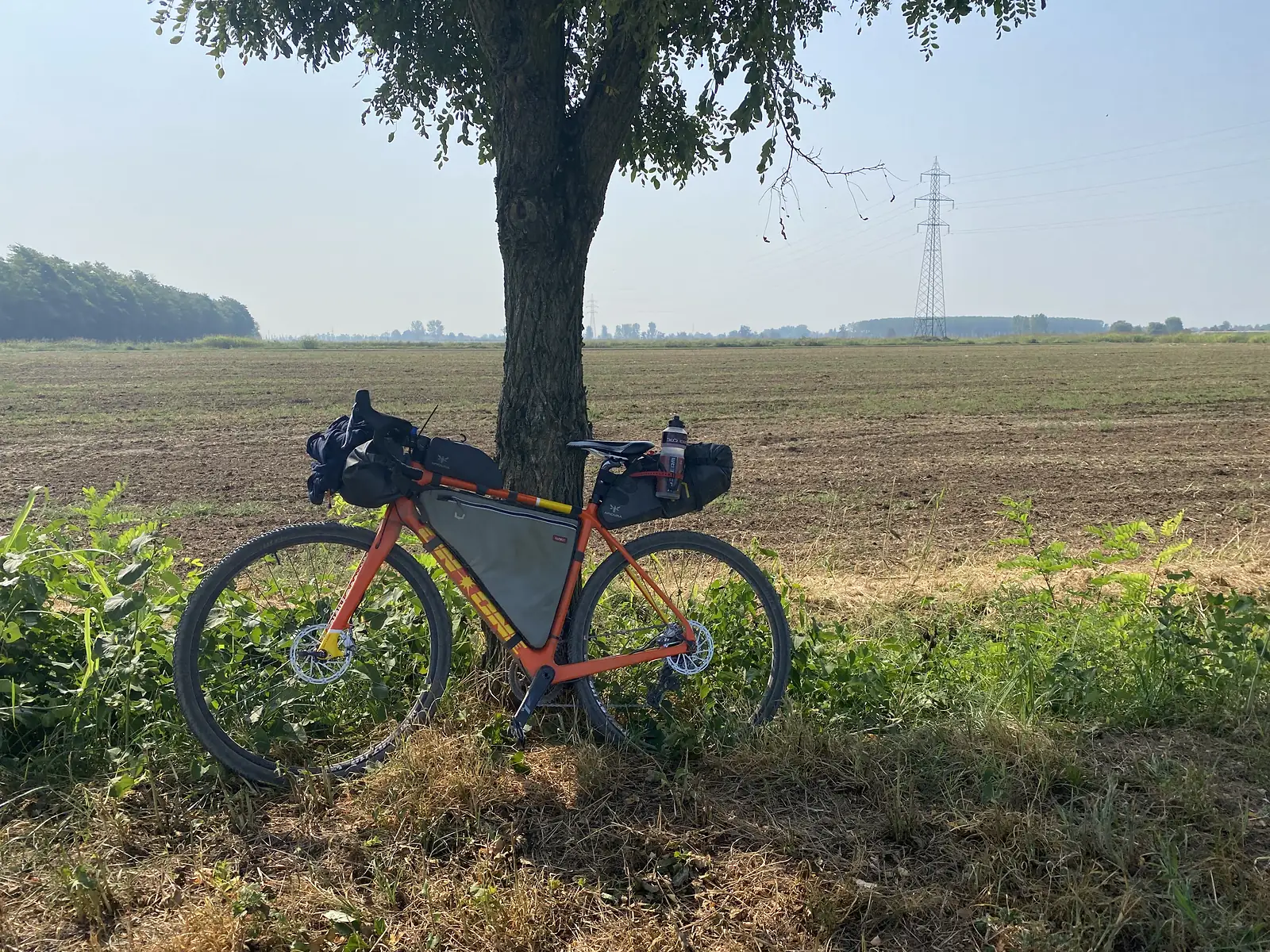 A laden orange gravel bike leans against a roadside tree, its frame bag and panniers strapped on, with a pylon and open fields behind.