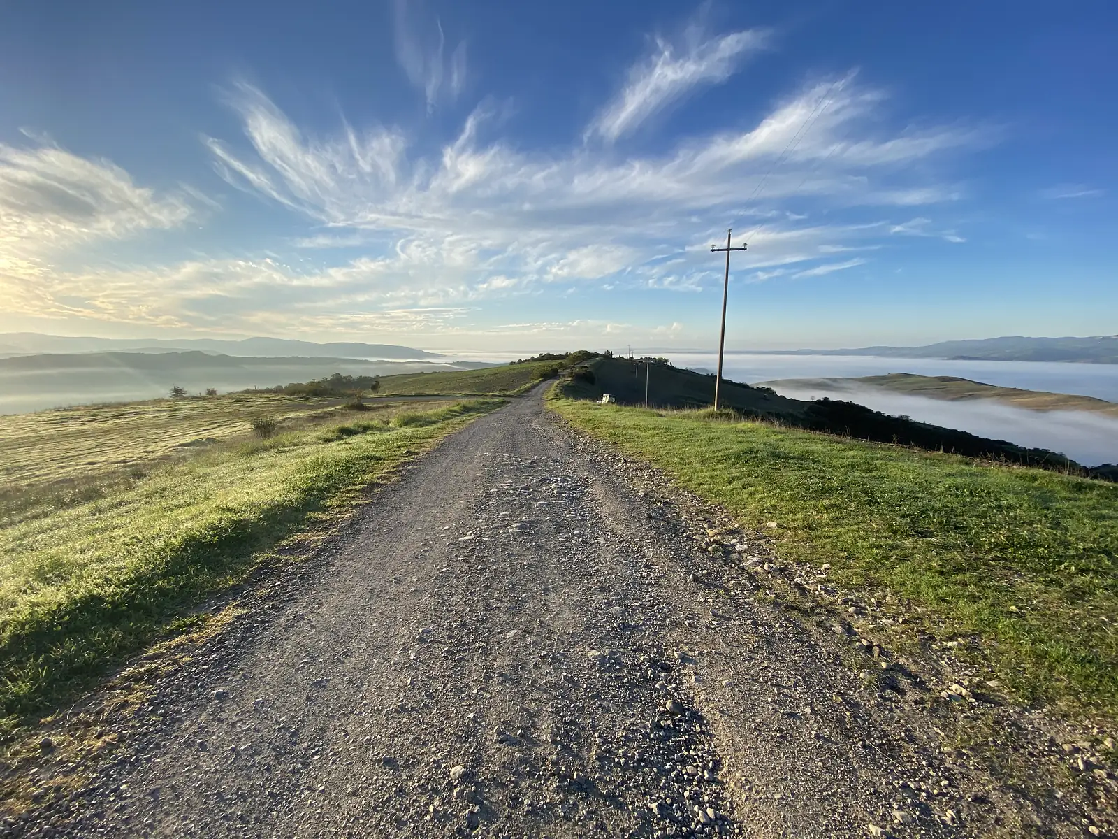 A gravel ridge-top track running into the distance with a wooden power-line pole beside it, a thick sea of cloud filling the lowlands on either side under a wispy blue sky.