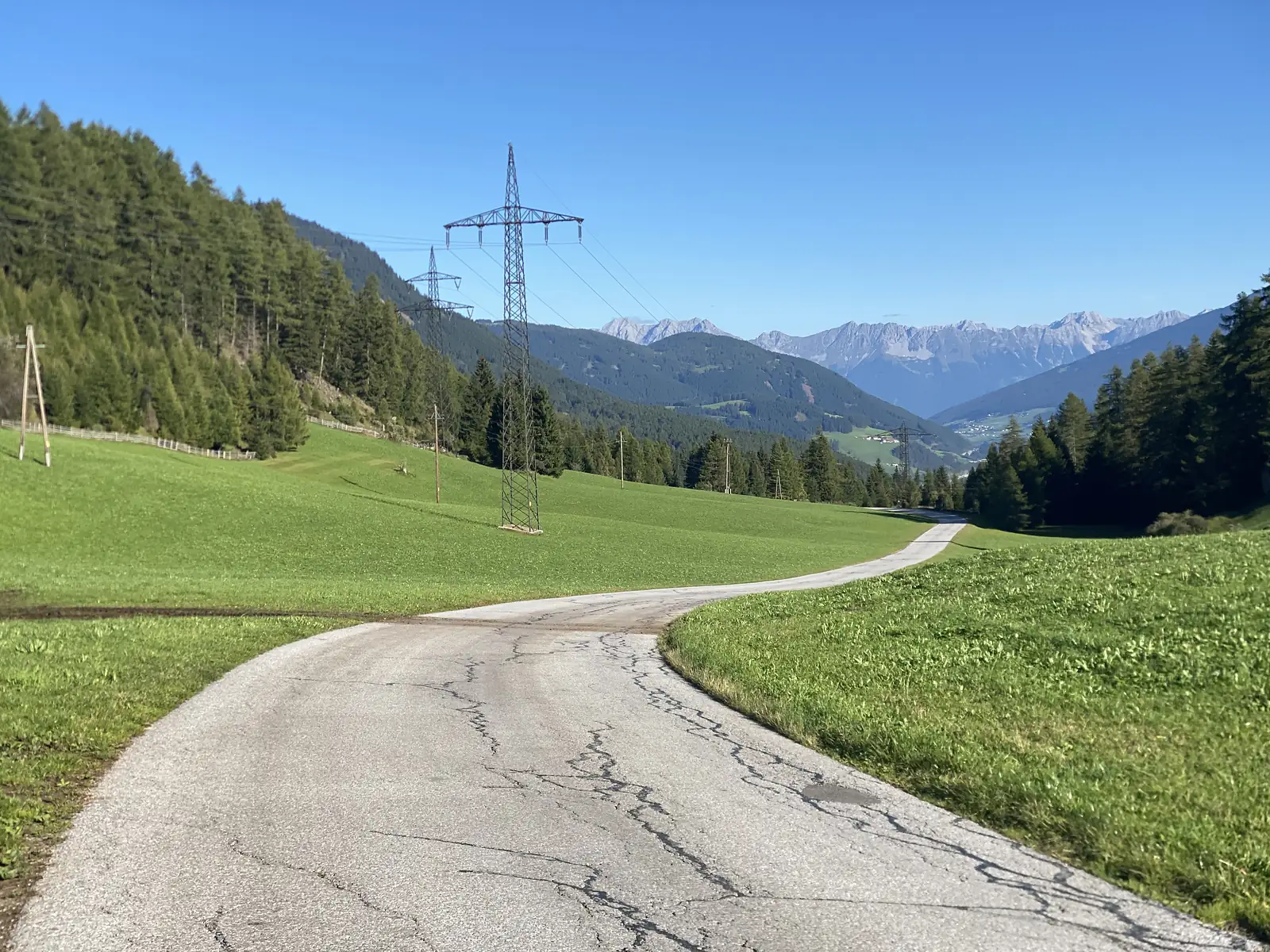 A cracked single-track lane runs down through mown meadows past an electricity pylon, with a long serrated mountain range on the horizon.
