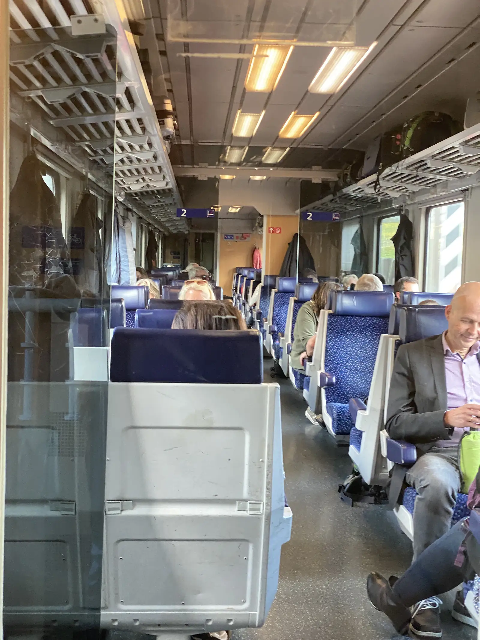 Passengers sit among luggage in a second-class train carriage with blue upholstered seats.